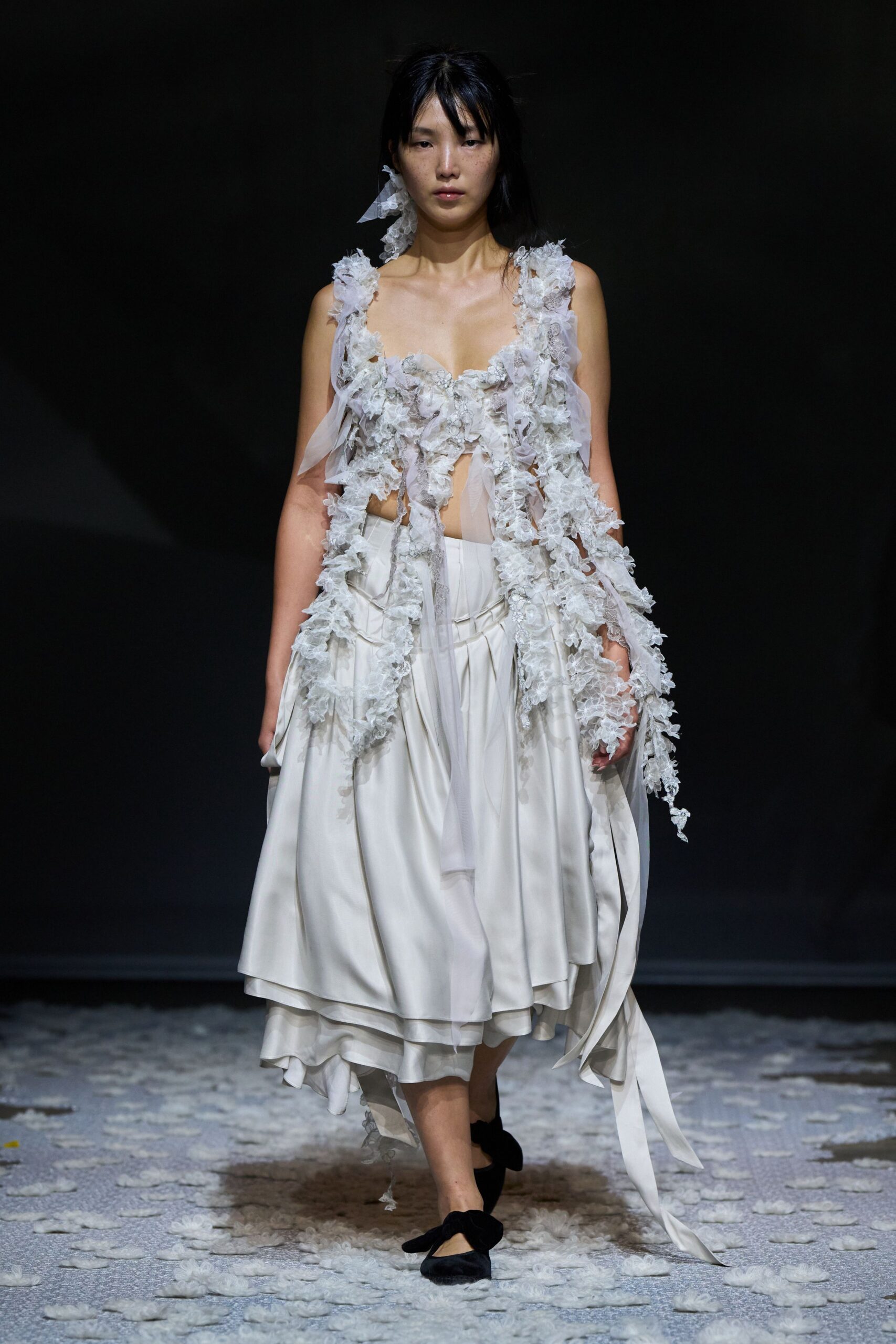 Ivory dress with a plunging bodice buried under shredded organza blossoms. Layered satin skirt, softly gathered, with a trailing chiffon tie. Black velvet flats with oversized bow fronts. Crossing a chapel floor after the petals have already been crushed.