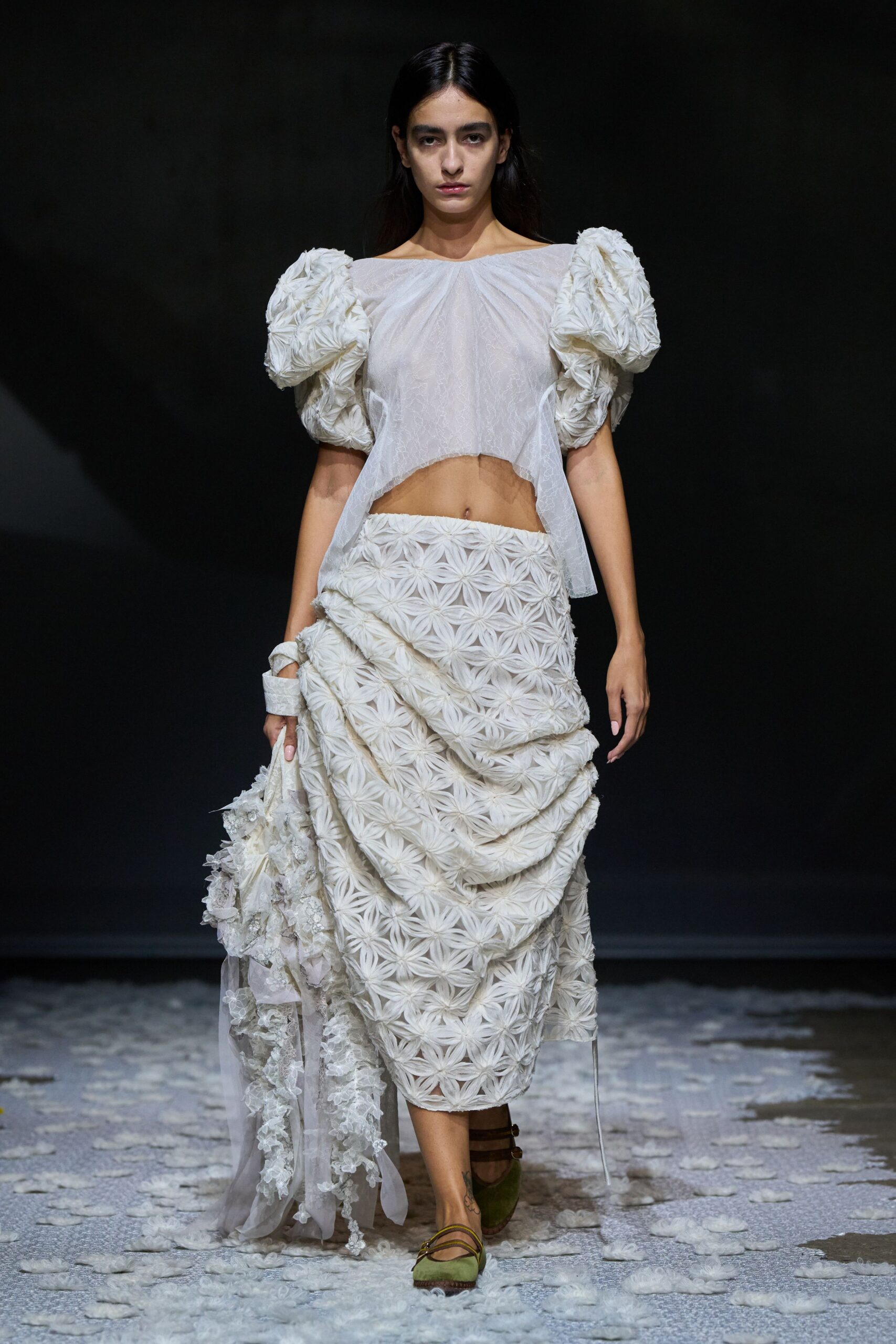 Sheer white gauze top, hem dipping to expose the navel. Puffed sleeves built from ruched rosette circles. Matching rosette skirt draped into heavy swags at one hip. Trailing lace bundle looped over the wrist. Olive flats with thin brown straps. Crossing a churchyard after the wedding, carrying what came loose.