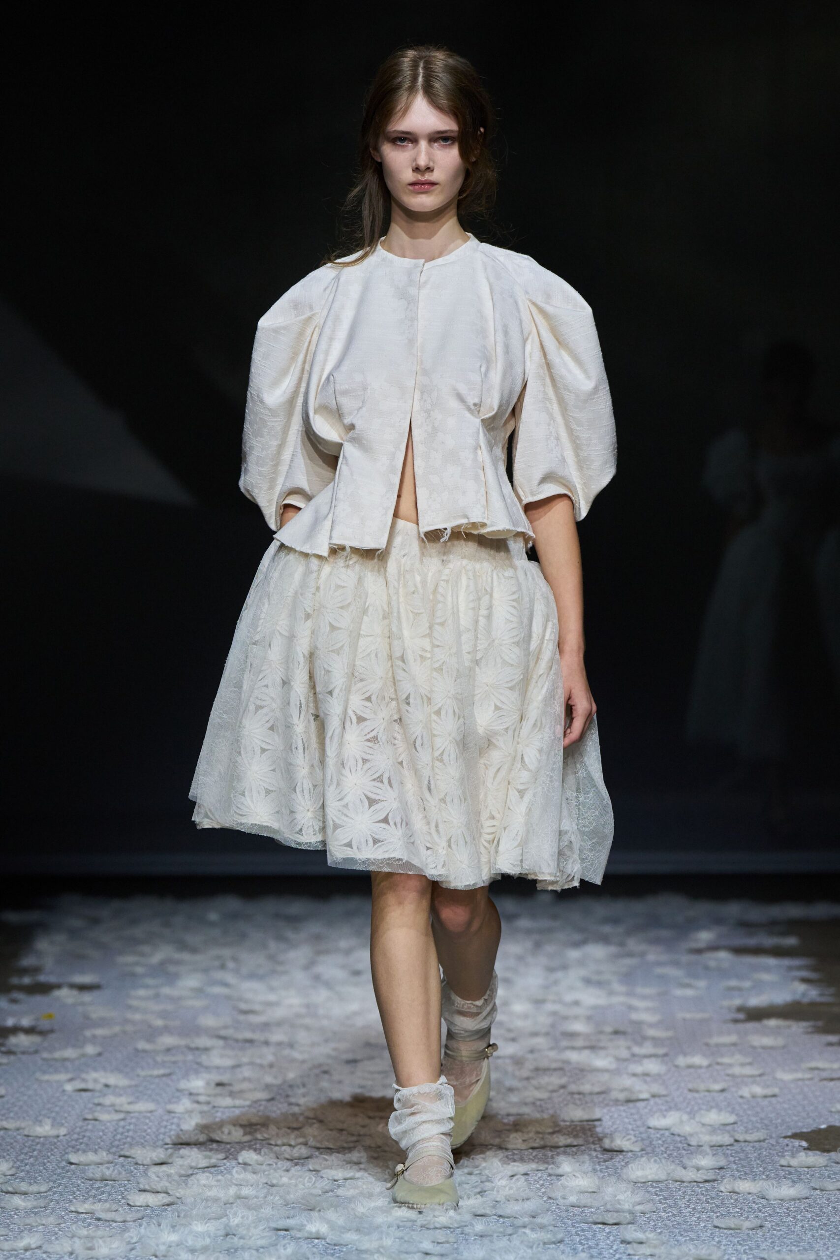 Ivory jacquard blouse with puffed sleeves and a frayed split hem. Full white skirt layered over petal-cut lace, softly clouded at the edge. Sheer socks, ruched and twisted. Pale flats with a narrow strap. Crossing a frostbitten conservatory after the wedding guests have gone.