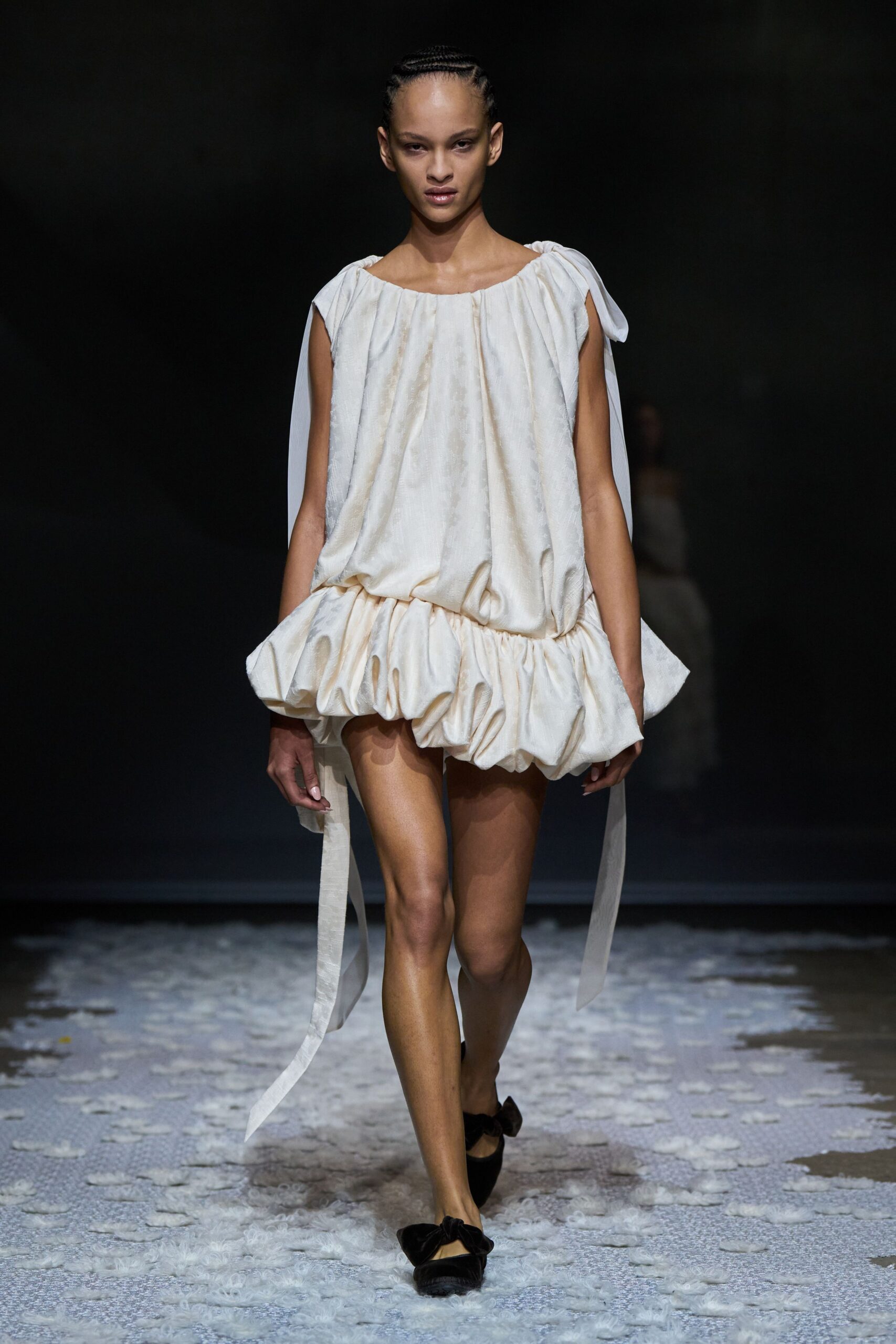 Ivory satin mini dress, gathered boat neck and inflated bubble hem. Long self ties falling from both shoulders, one knot slightly heavier. Black velvet flats with oversized bow vamps. Morning after the wedding, crossing a cold hotel lawn barefoot in your head.