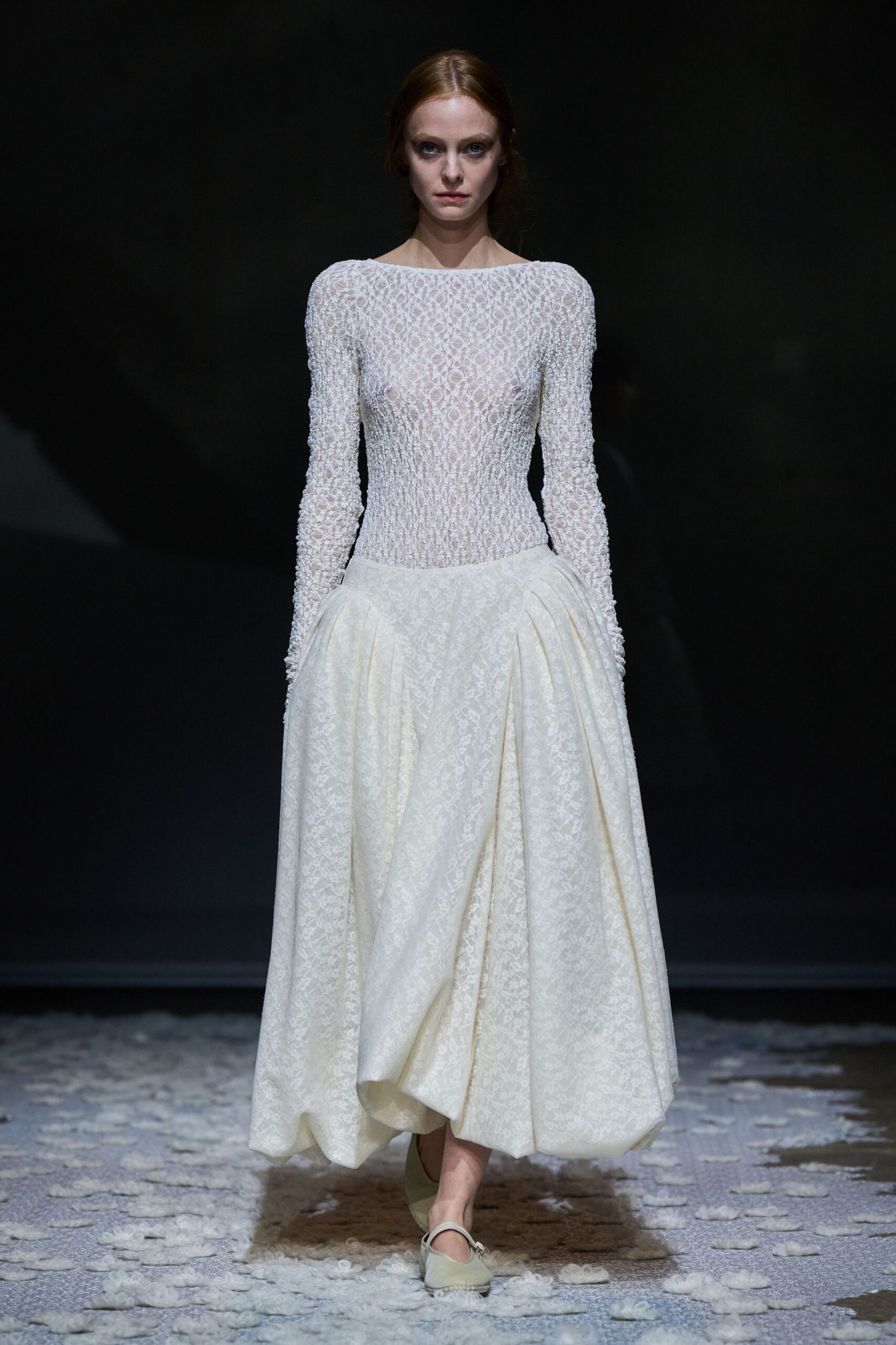 Ivory boat-neck dress, sheer looped lace bodice pulled tight through the waist. Dropped-waist skirt in pale floral jacquard, gathered into deep side swags. Cream Mary Jane flats with a slim strap across the instep. Crossing a cold chapel floor after everyone else has gone outside.