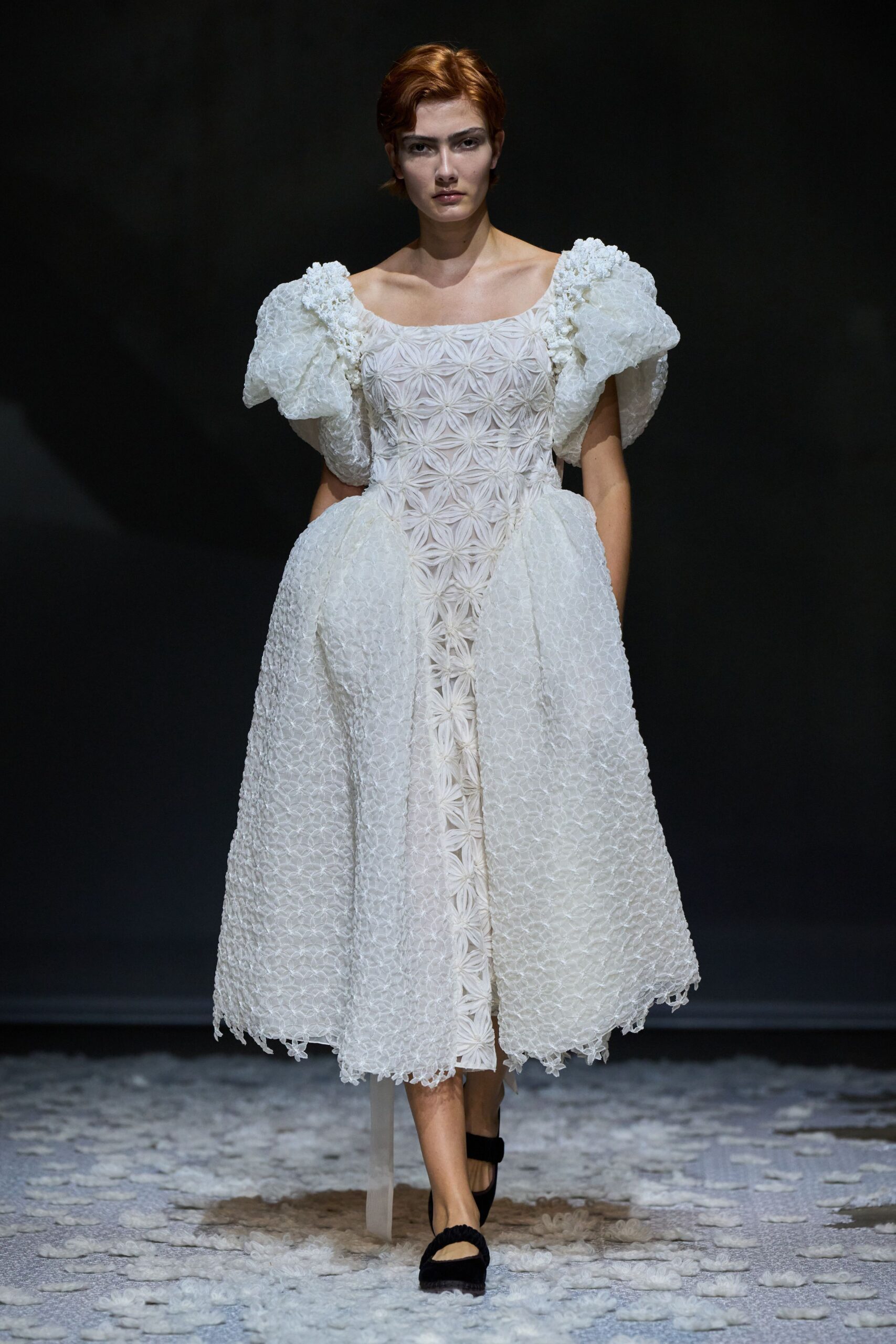 White floral-smocked dress with balloon sleeves and padded hips jutting outward. Black Mary Jane flats with a softly ruched instep strap. Crossing a chapel aisle after the rice has already hit the floor.