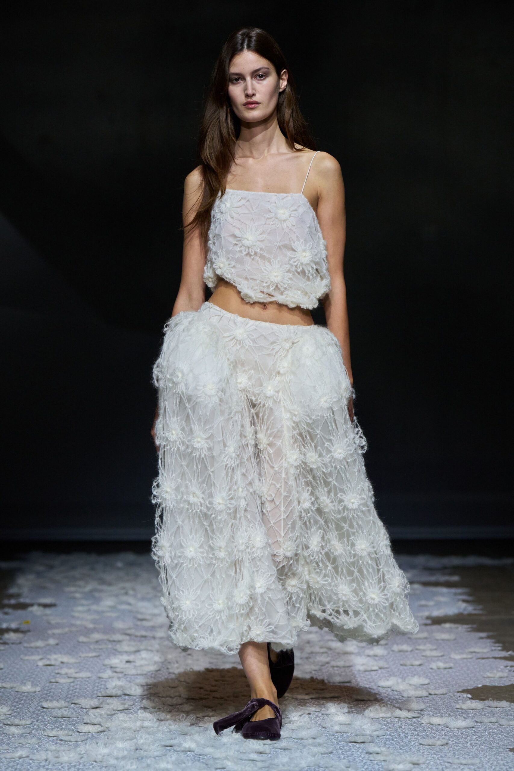 Sheer white camisole in floral looped lace, hem puffing softly above the navel. Matching calf-length skirt with airy raised daisies and a clouded overskirt volume. Deep plum velvet flats with a twisted strap crossing the instep. Crossing a hotel lawn after midnight, petals stuck to damp paving.