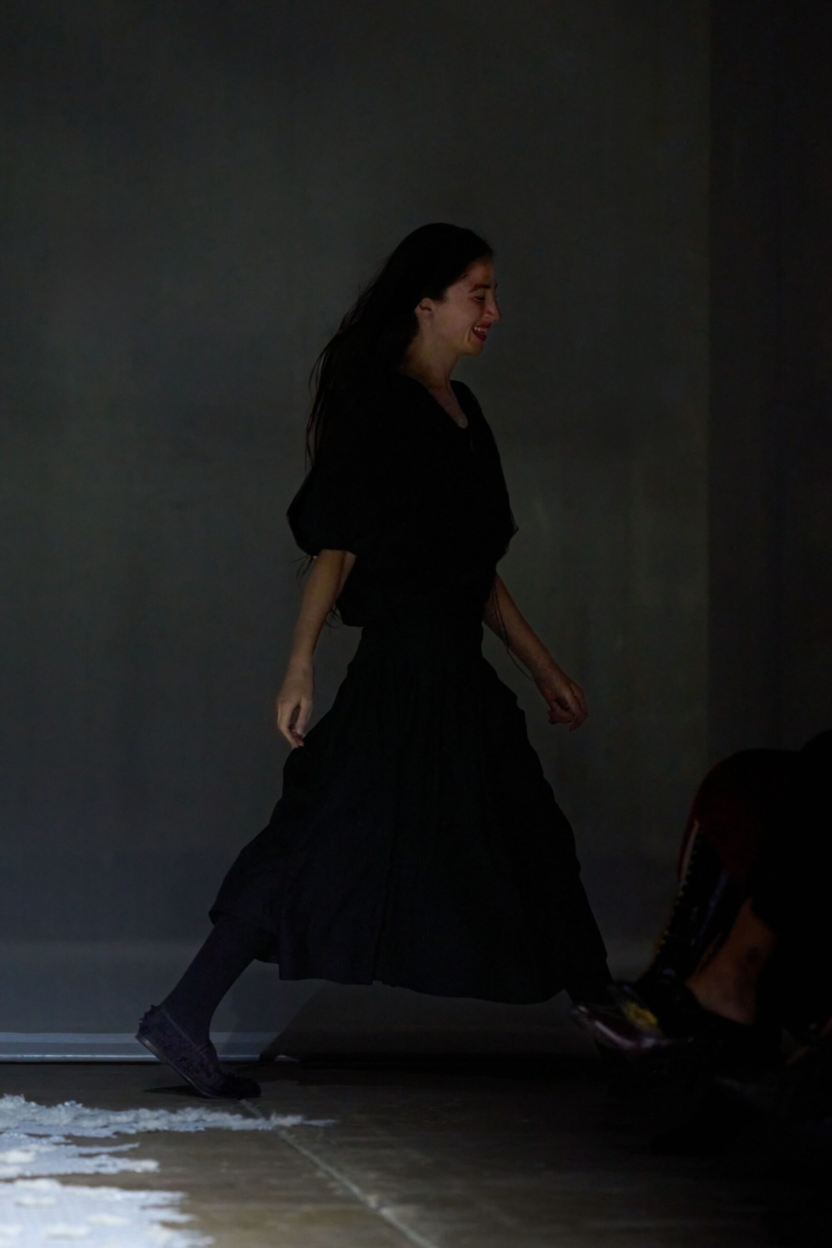 Black matte dress with dropped shoulders and a densely gathered midi skirt. Opaque black tights. Soft black lace-ups, one untidy bow end trailing. Crossing a church hall floor after the folding chairs have been stacked.
