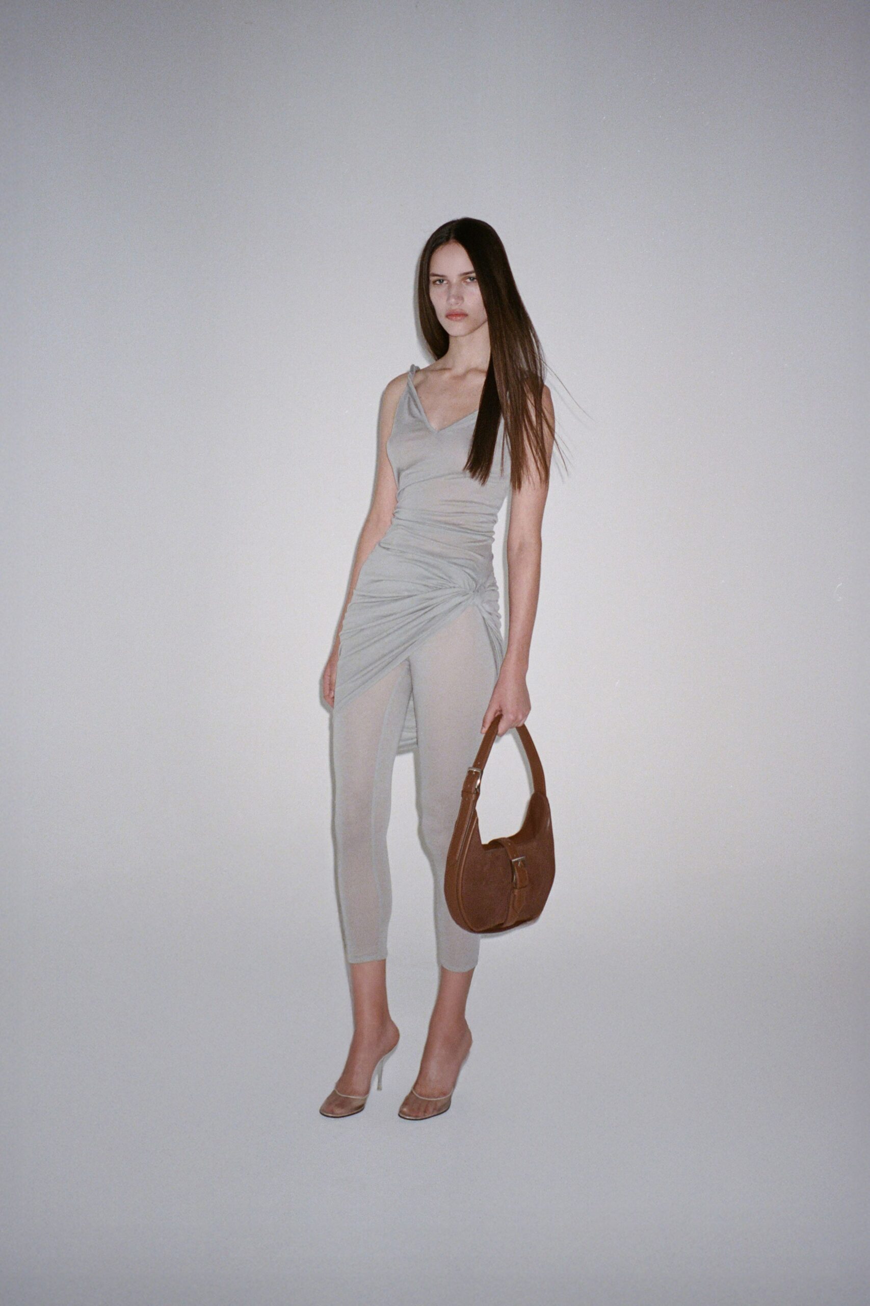 Pale dove jersey dress, spaghetti straps and a twisted hip drape. Matching cropped leggings disappearing under the dress's gathered front. Chestnut crescent shoulder bag with a buckle-tab pocket. Clear-strap mules on slim white heels. For crossing a hotel lobby barefoot in your head, heels still on.