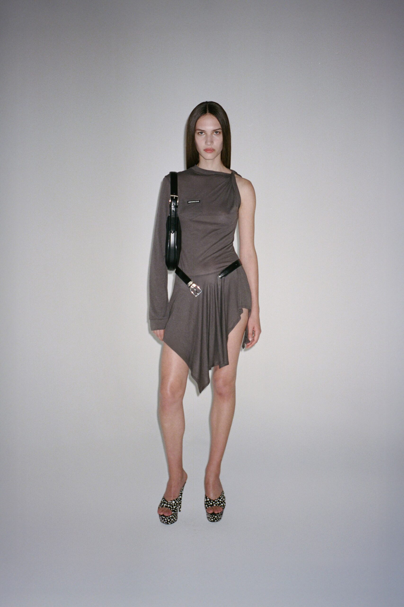 Taupe jersey one-sleeve mini, twisted at the neck and hip. Black leather belt slung low, silver buckle landing off-center. Slim black shoulder bag tucked tight under one arm. Black mule sandals crusted with silver studs. Waiting for the hotel lift after midnight, keycard gone missing again.