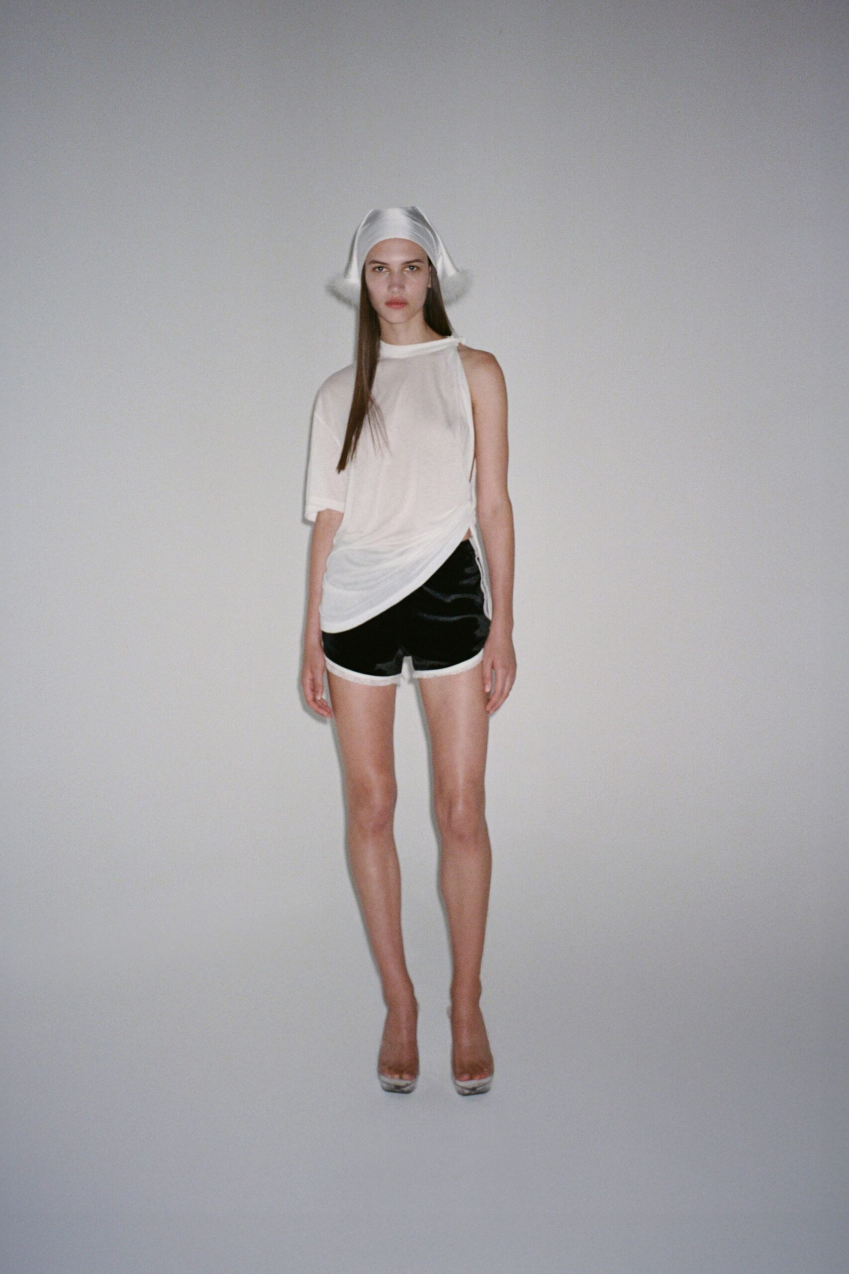 White satin headscarf with soft fur puffs at each side. Sheer white jersey top, one sleeve dropped and hem twisted low. Black satin track shorts with white binding at the rolled edge. Clear platform mules. Walking back from the hotel pool after midnight, still carrying chlorine.