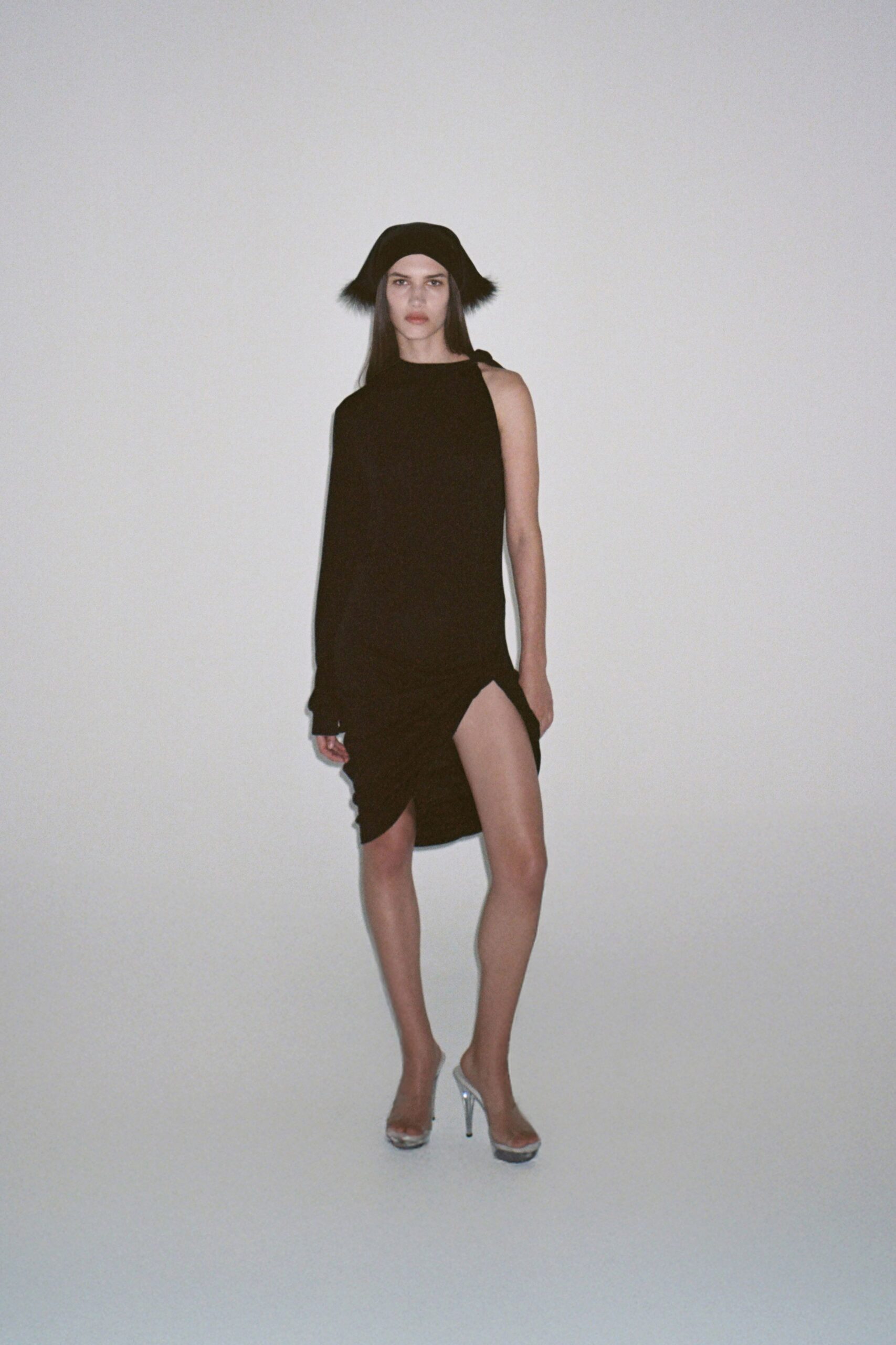 Black felt hat with short fur tufts kicking out at the sides. Black draped dress, one sleeve missing and hem twisted into a thigh slit. Clear vinyl mules on thin silver stiletto heels. Whatever you pull on for the hotel corridor ice machine at 2 a.m.