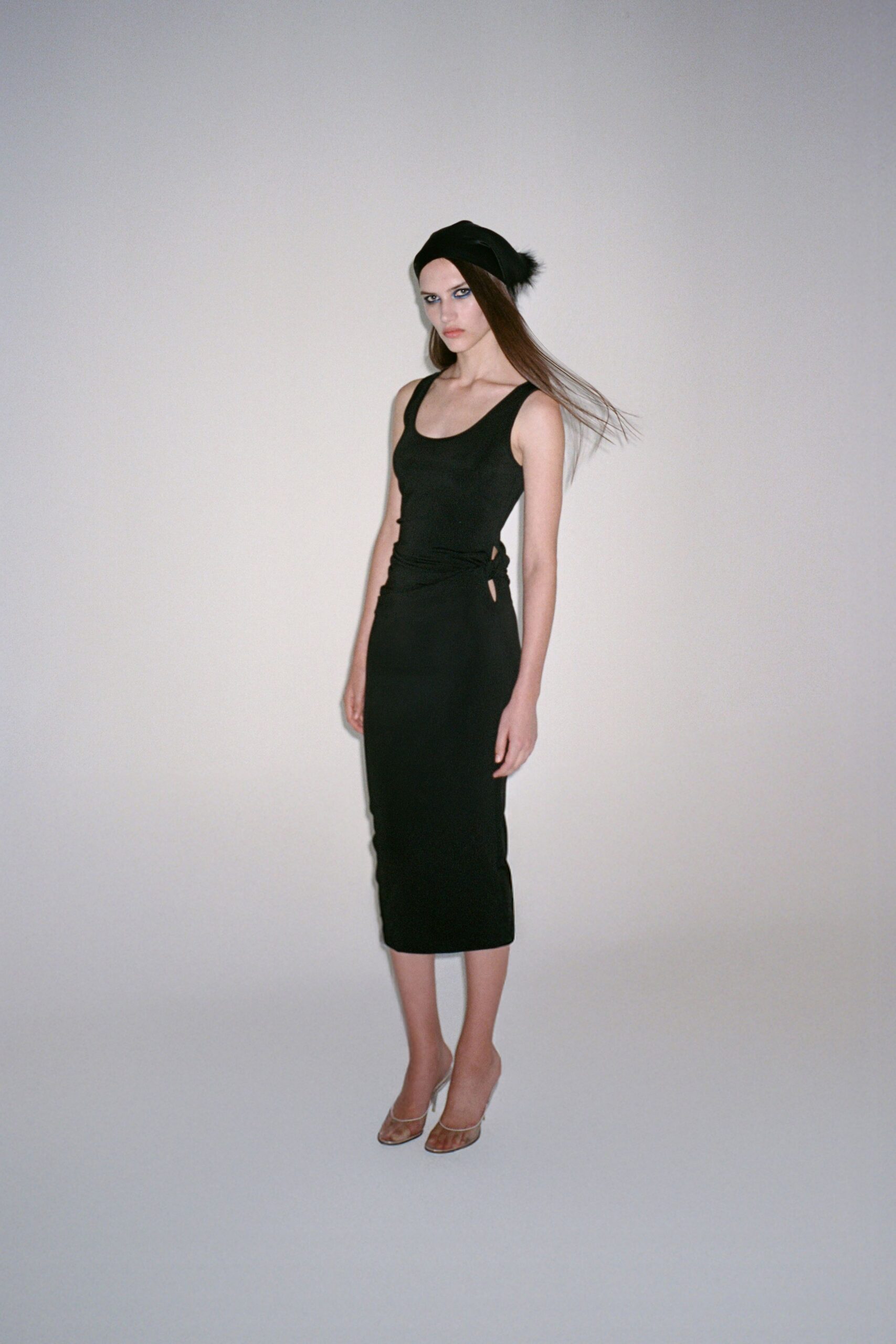 Black knit beret with a small fur tuft at one side. Black jersey tank dress, side waist knotted through a narrow cutout. Clear-strap heeled mules with a squared toe. For the walk home after midnight, carrying your shoes by the straps.
