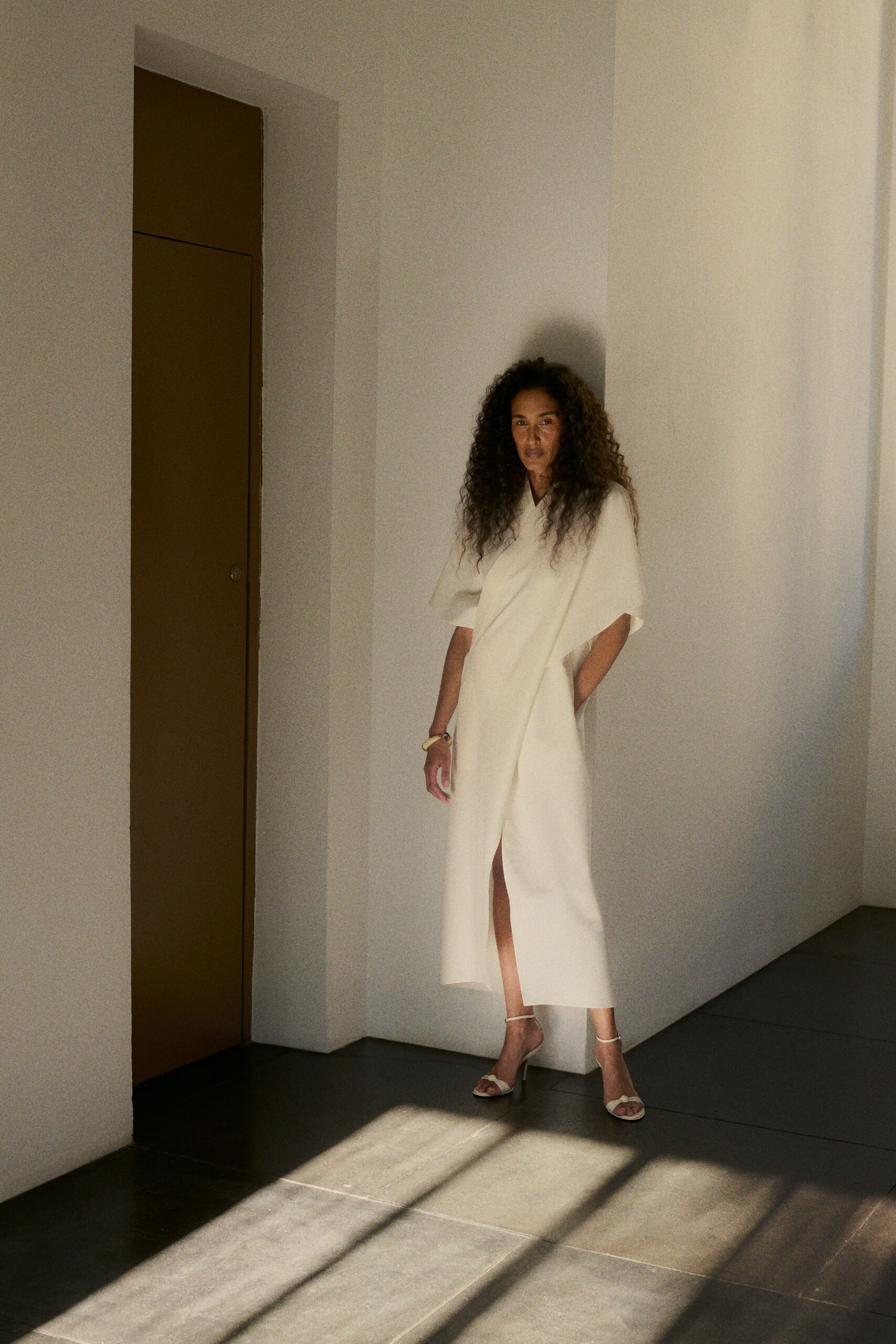 Ivory wrap dress in dense crepe, one sleeve falling to a cape panel. Chunky pale resin bangle. White ankle-strap sandals with a twisted toe band. Afternoon heat in a borrowed villa, standing where the cool tile begins.