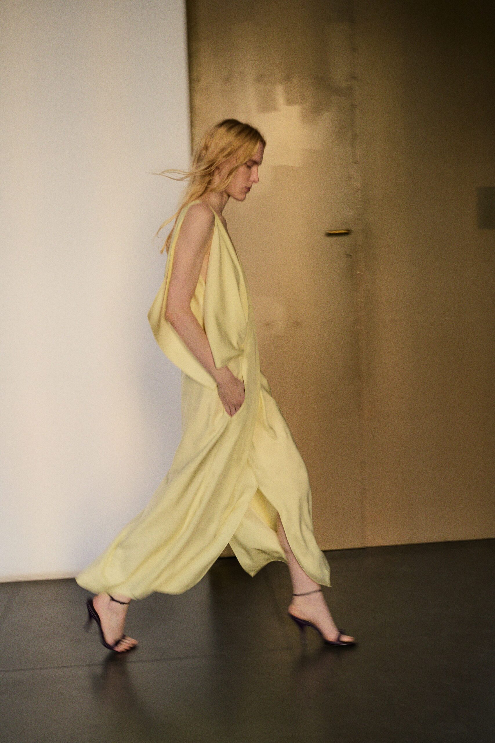 Butter-yellow draped dress in fluid crepe, deep armholes and pocketed folds. Black ankle-strap sandals with a narrow curved heel. Crossing a half-empty hotel corridor before breakfast service starts.