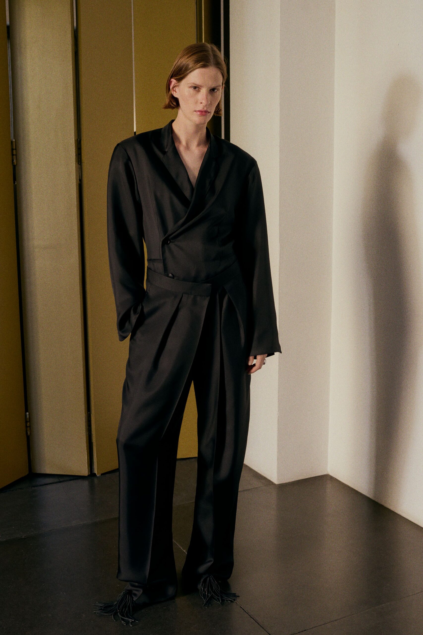 Black wrap-front jacket in matte suiting, one button set off-center. Matching high-waisted trousers with a folded waistband crossing the hip. Black shoes finished with long leather fringe over the toes. For crossing a hotel lobby before sunrise with your keycard already in hand.