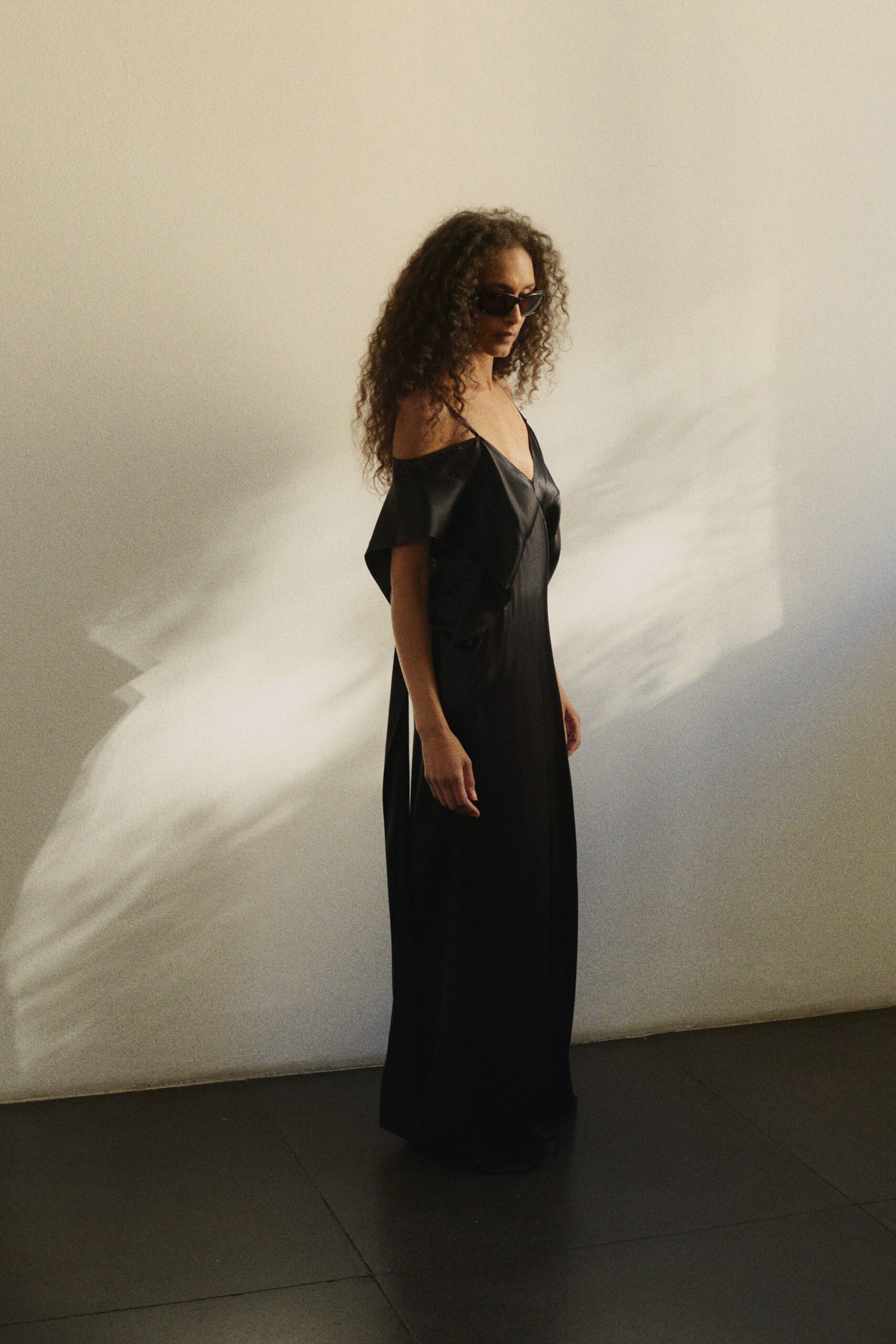 Narrow black sunglasses with sharply upturned corners. Black satin column gown, camisole straps and fallen cape sleeves framing the chest. Sun through museum blinds, long after the opening remarks ended.