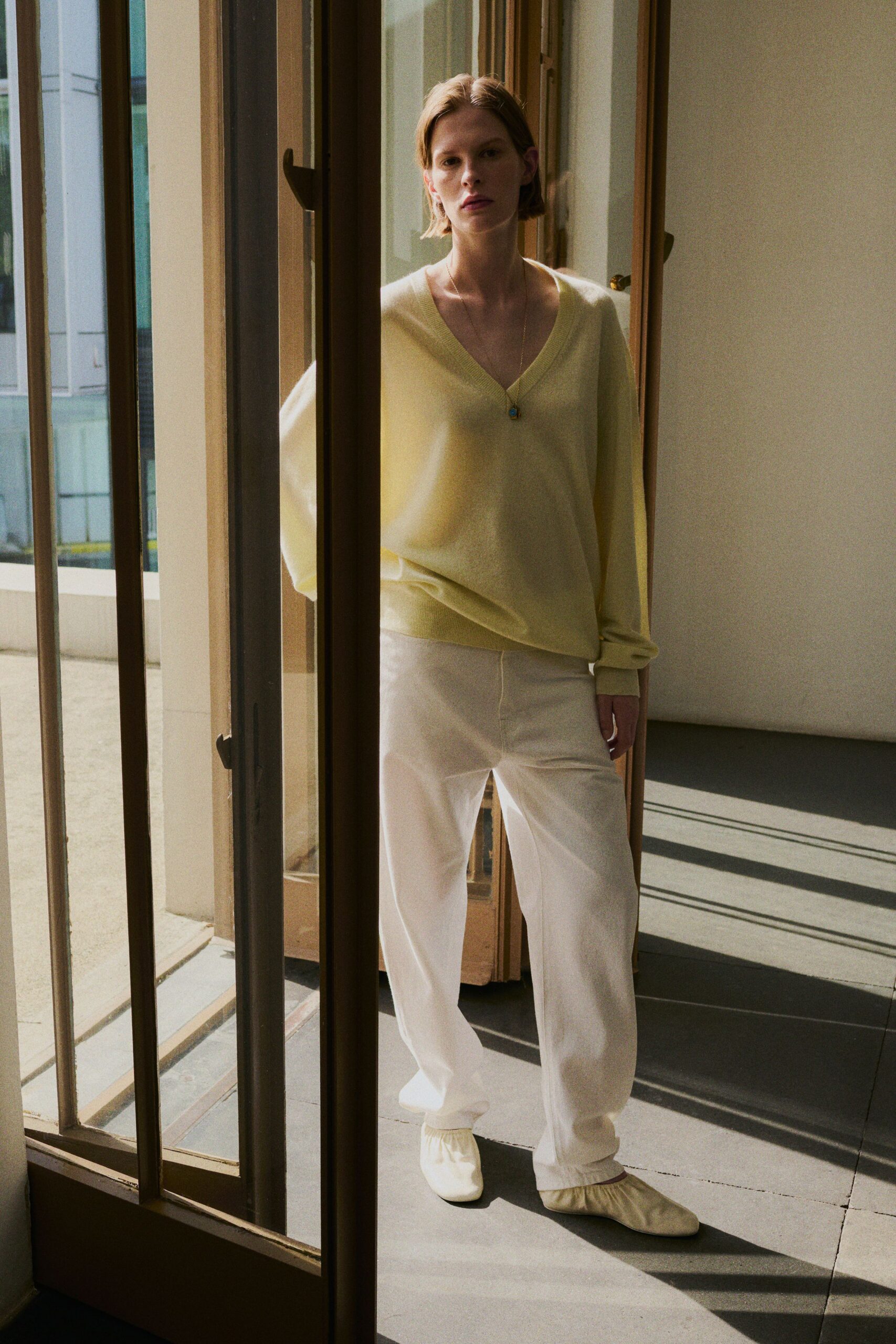 Pale citron fine-knit V-neck sweater, slouched wide at the ribbed hem. Long chain with a tiny turquoise pendant at the sternum. White drawstring trousers in soft twill, pooling lightly over the feet. Cream gathered flats with a ruched opening. Sun through open doors, borrowing somebody else's apartment for the afternoon.
