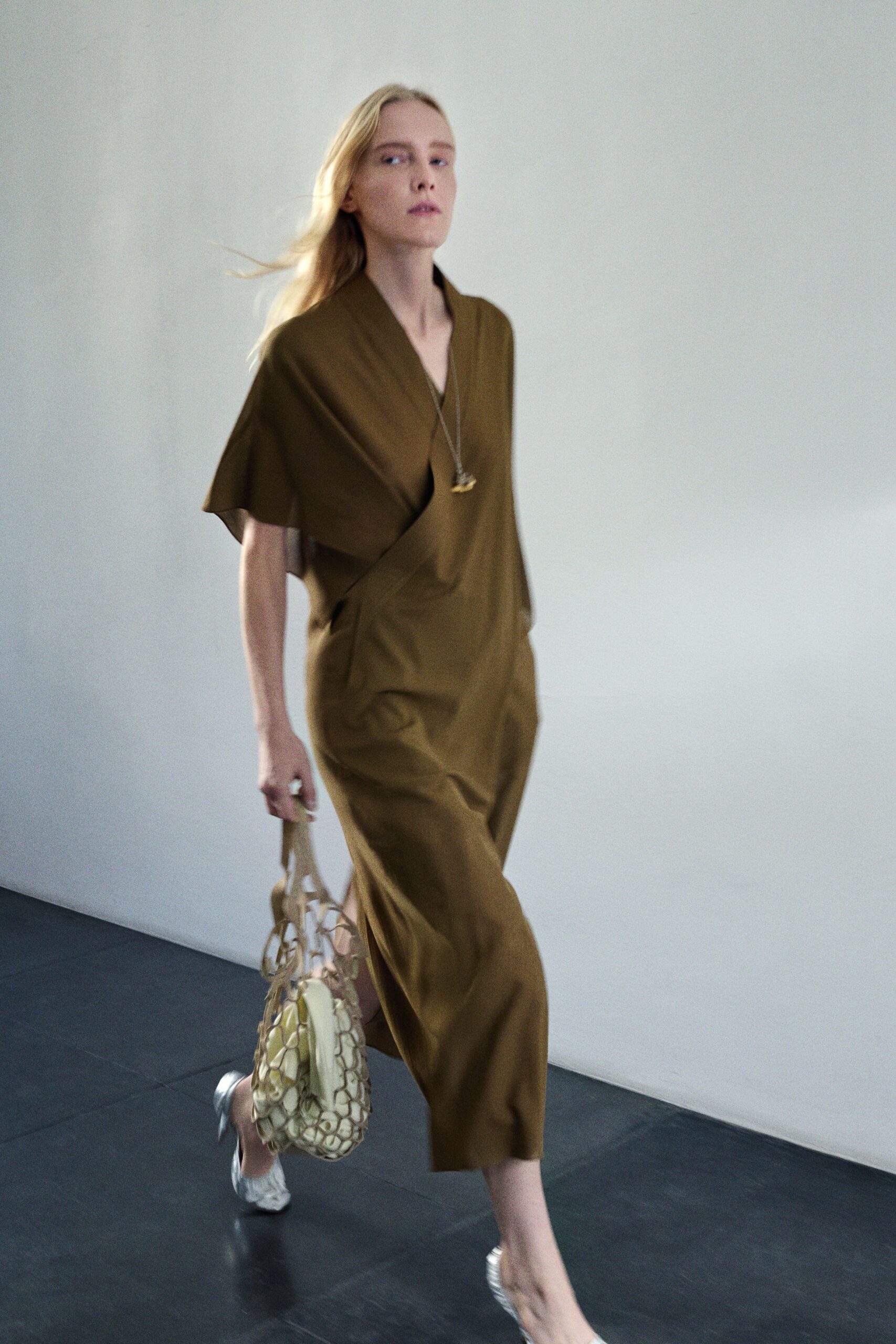Olive draped dress in fluid twill, one cape sleeve falling from the shoulder. Long chain with a small gold pendant at the sternum. Cream netted bag stuffed with pale rounded forms. Silver flats, softly crushed at the toe. Crossing a museum lobby with flowers and fruit for someone's new apartment.