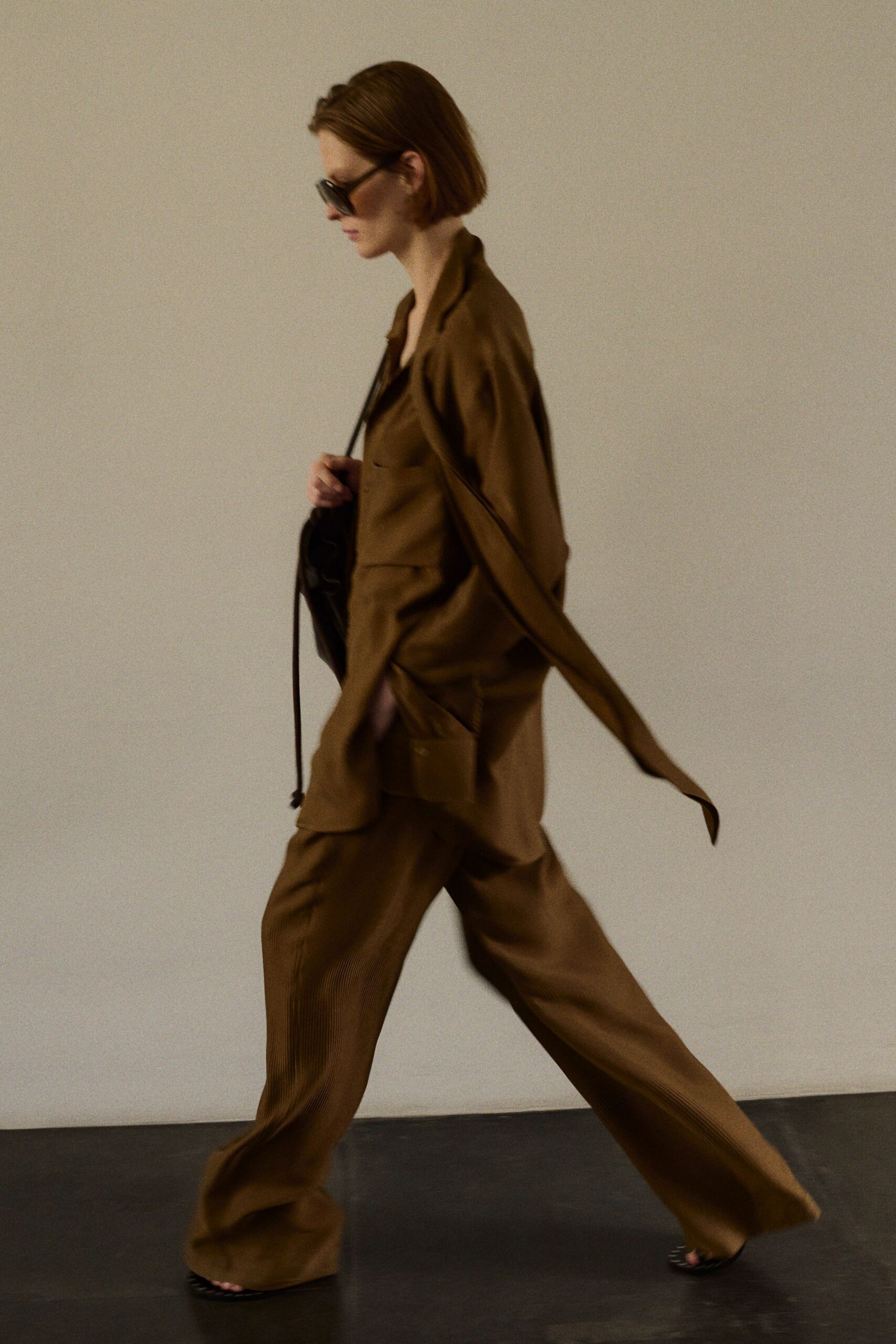 Oversized black acetate frames hiding the eyes. Tobacco draped jacket with a long self-tie streaming behind. Dark liquid-sheen camisole cut low at the chest. Wide tobacco trousers in fine vertical pleats, pooling over the feet. Black toe-baring sandals. Crossing a near-empty lobby before the air conditioning fully wakes up.