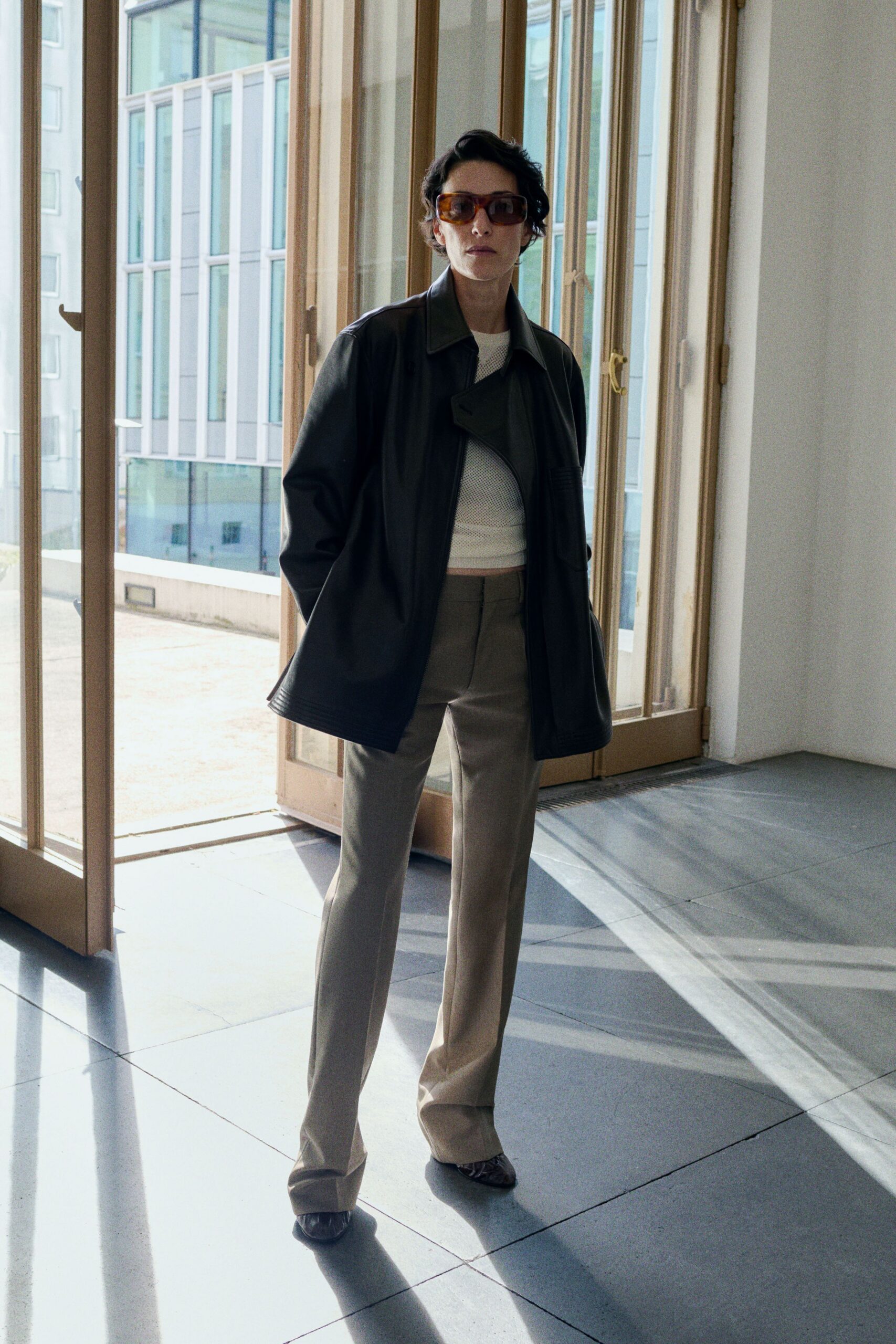 Amber shield sunglasses with thick squared rims. Black leather jacket cut long, one lapel folded across the zip. White mesh crop top over a pale underlayer. Taupe trousers with a pressed crease and long break. Dark snakeskin pumps, sharply pointed. Sun through office windows after everyone else has left for lunch.