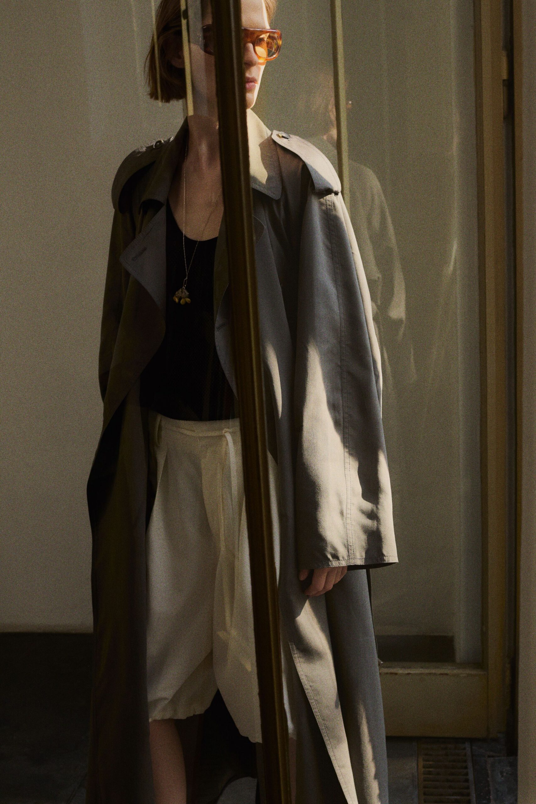 Amber-tinted wraparound lenses. Stone cotton trench, epaulettes buttoned and lapels left to fall open. Black scoop-neck tank in a matte jersey. Long chain with three small yellow bead drops. White pleated shorts with a folded paperbag waist. Sun coming through office blinds after a lunch that ran too long.
