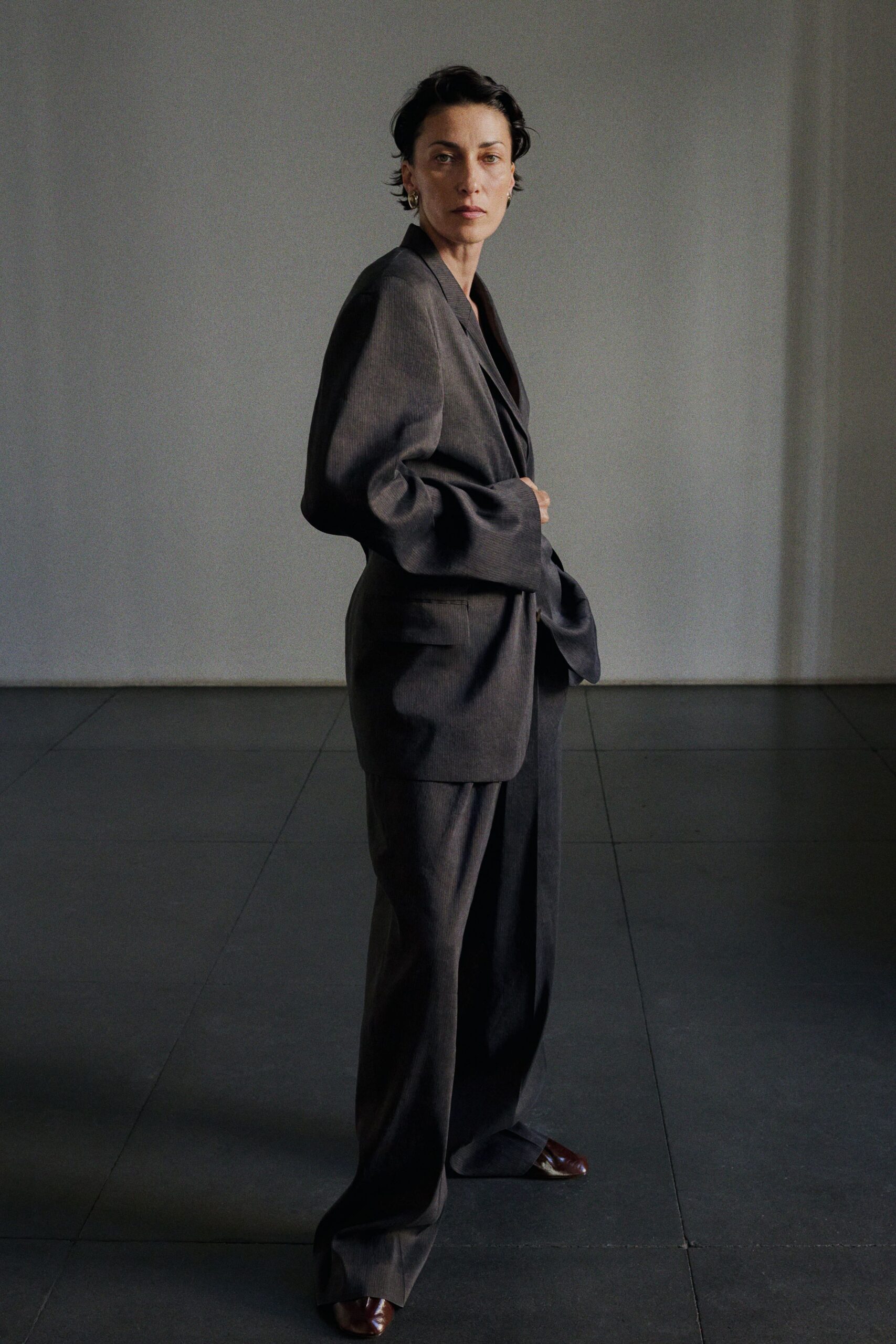 Charcoal worsted blazer cut broad through the shoulder, one hand holding it closed. Matching loose trousers breaking deep over the shoes. Oxblood leather square toes peeking from the hem. Small gold hoops. For the first meeting in a borrowed office that still smells of fresh paint.