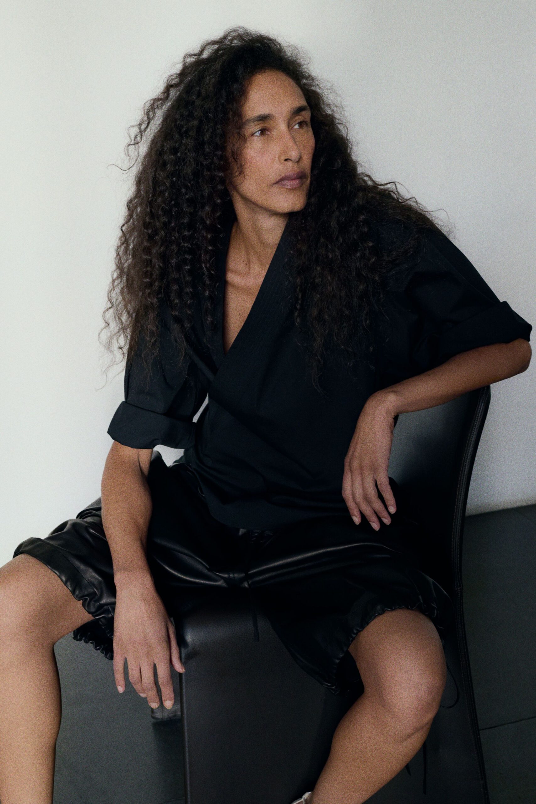 Black wrap top with rolled sleeves and a fine pinstriped lapel. Black leather shorts, gathered hem puffing lightly around the thigh. For the first meeting after lunch, blinds half closed against the heat.