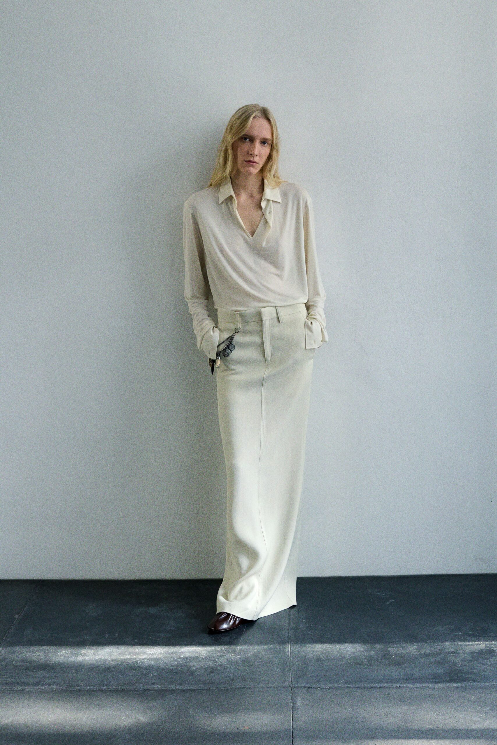 Ivory satin blouse with a soft drape and deep open placket. Floor-length ivory column skirt, pressed center seam falling almost to the toe. Gunmetal chain clipped at the hip with a dangling dark charm. Dark brown leather square toes peeking from the hem. Morning in an empty apartment before the movers bring everything back.