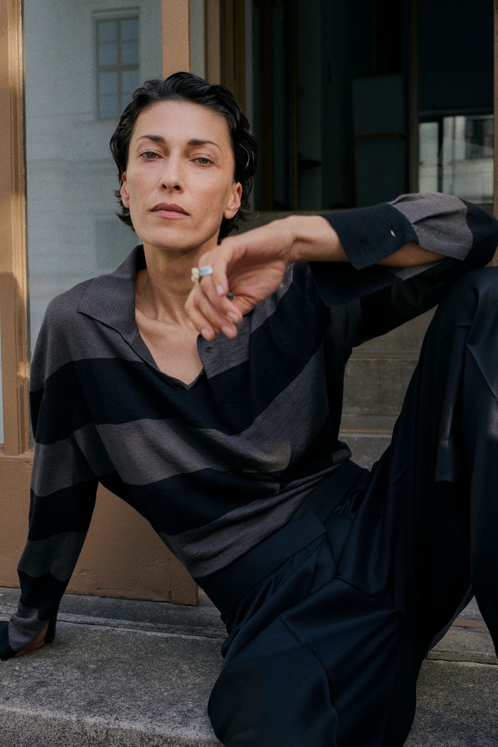 Charcoal and black rugby-striped knit polo, placket left slack at the throat. Ink-dark wide trousers in fluid cloth, pressed folds breaking over the knee. Pale blue stone ring. On the stone steps after lunch, cigarette gone out between two fingers.