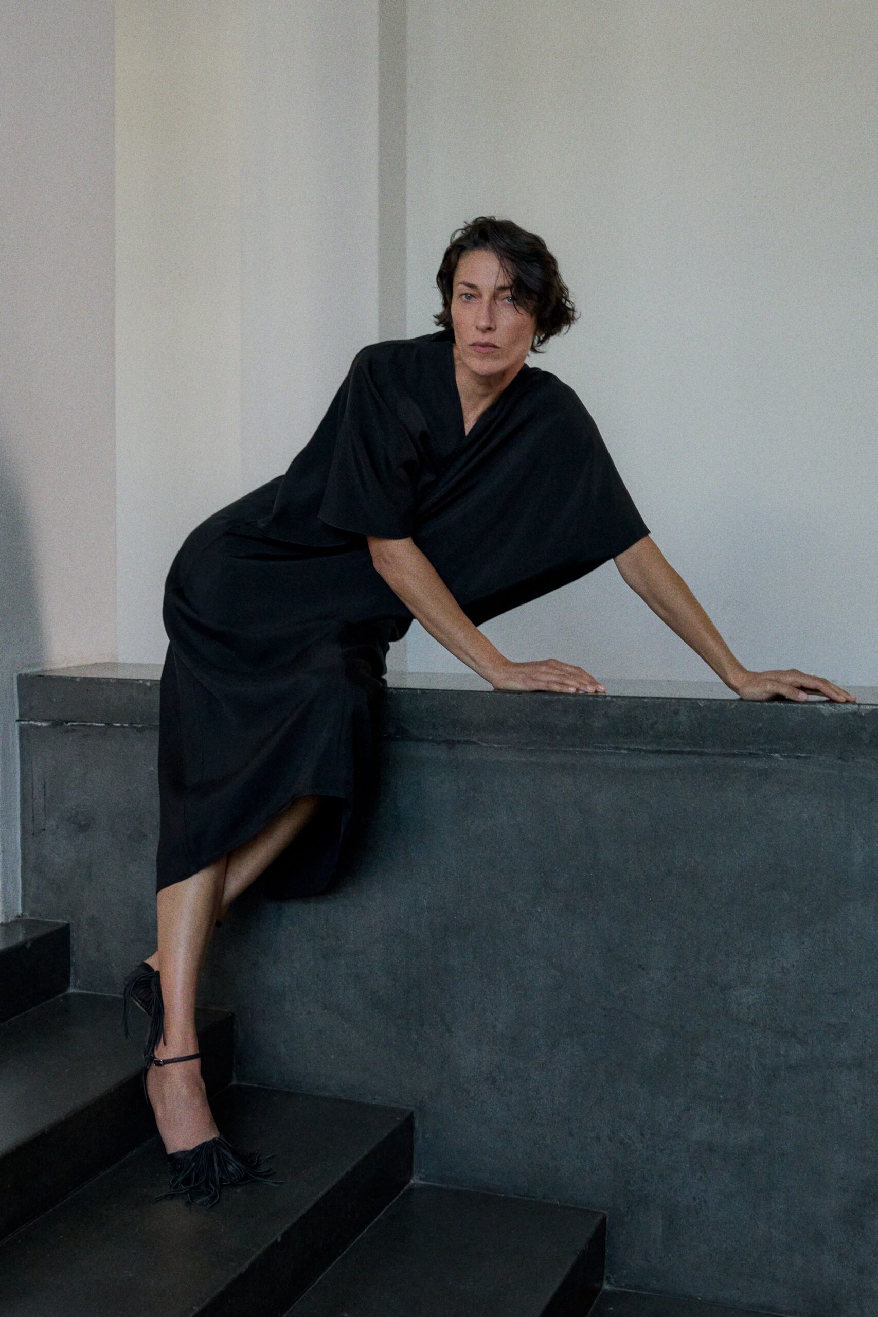 Black wrap dress in fluid matte crepe, one wide sleeve hanging almost square. Black ankle-strap heels with dense fringe bursting over the toe. Leaning across a concrete stairwell after everyone else has taken the lift.