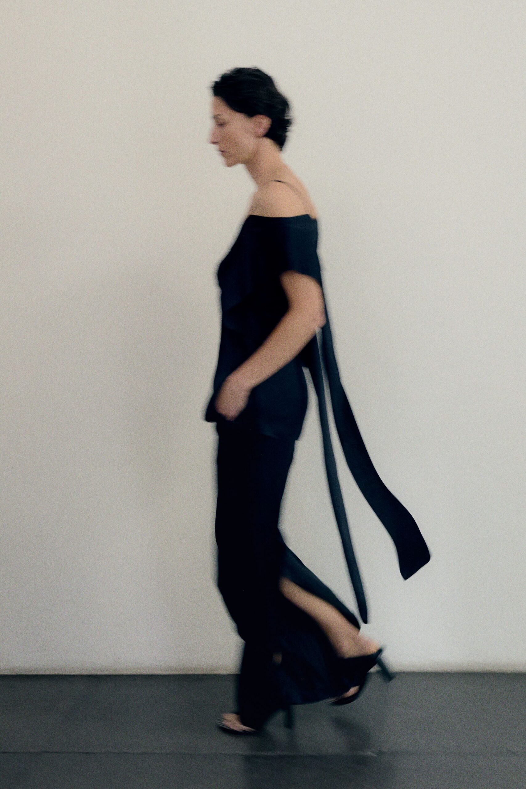 Black satin camisole top, one shoulder slipped low with trailing back ties. Black fluid trousers, hem opening into a calf-high slit. Barely-there black heeled mules. Crossing a hotel corridor barefoot in everything but the shoes.