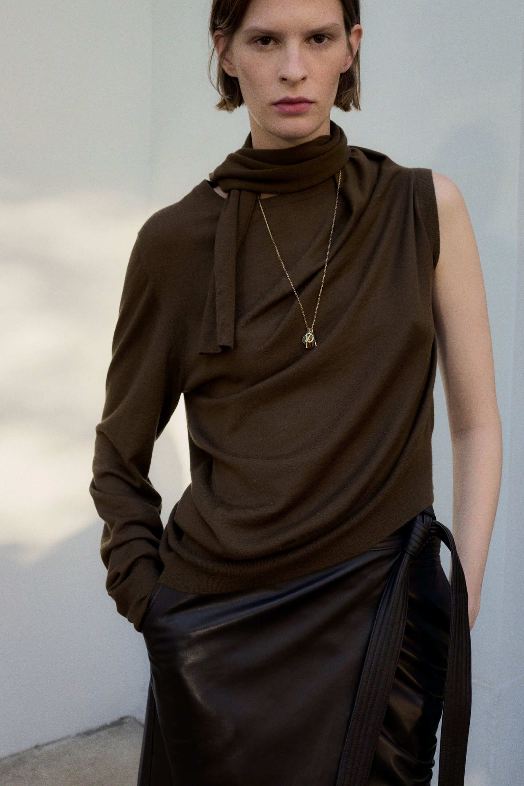 Espresso fine-knit top draped diagonally, one sleeve missing and scarf built in. Long gold chain with two tiny charms resting at the sternum. Black leather wrap skirt with a padded tie hanging past the hip. Waiting out drizzle under a museum portico after the offices emptied.