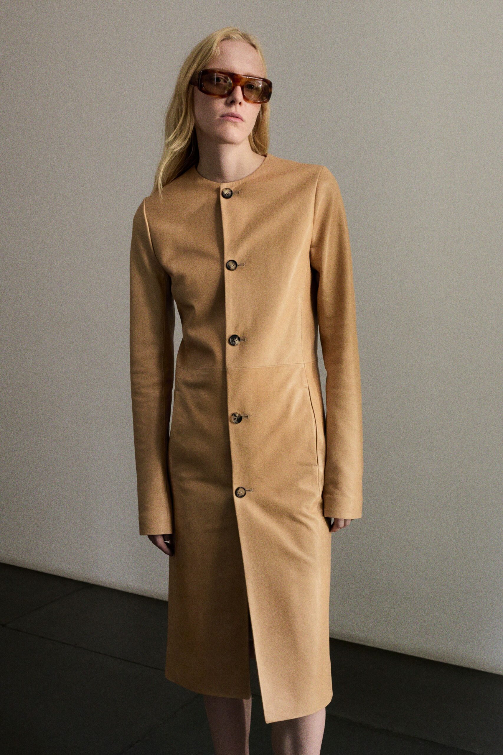 Oversized amber acetate frames with a squared-off wrap shape. Camel leather coatdress, collarless and spare, fastened with five horn buttons. Museum lobby at opening hour, heat still trapped in the stone.