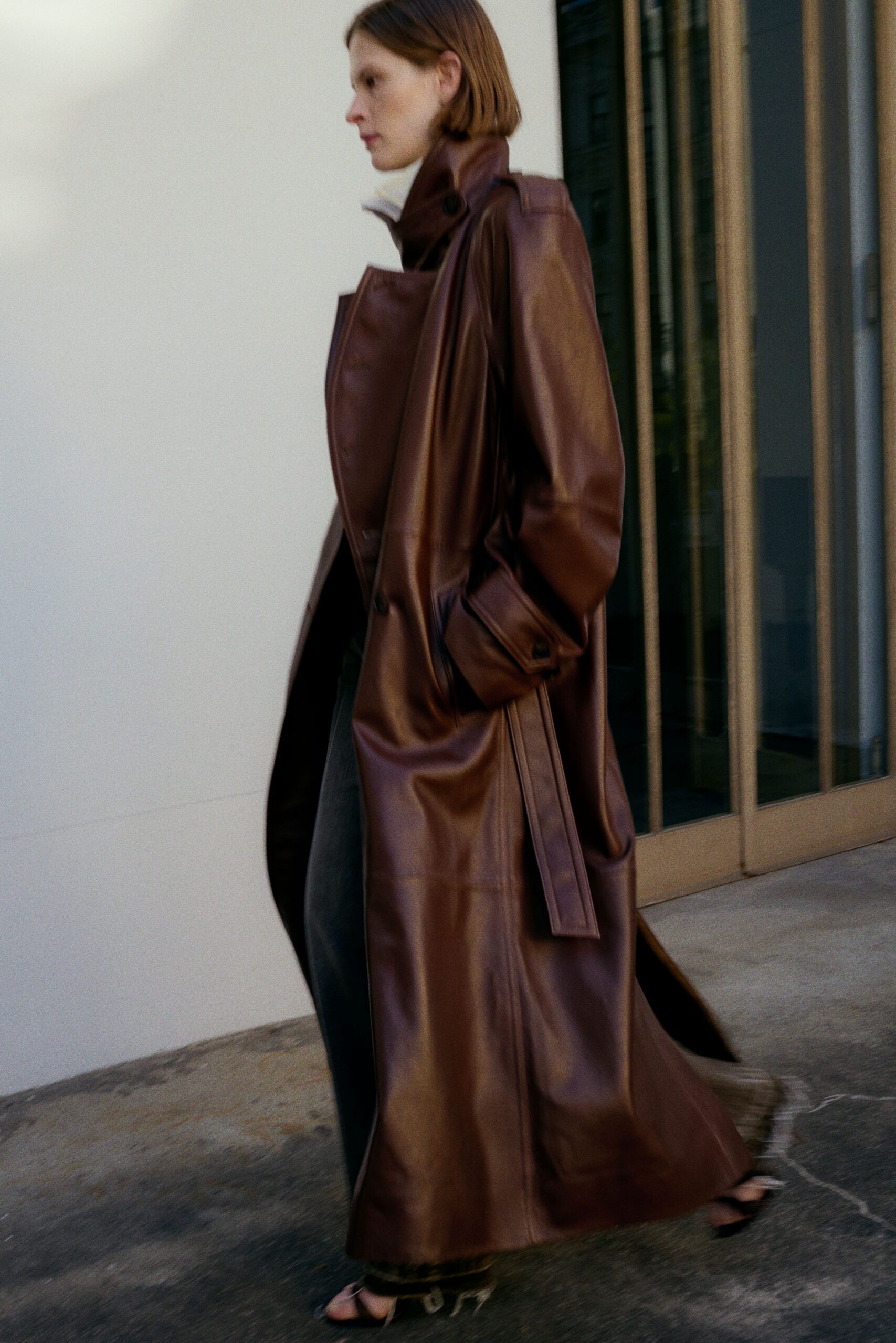 Mahogany leather trench, storm flap skewed across a high throat latch. Black leather trousers falling straight under the coat. Bare black thong sandals with a small metal ring at the heel. Crossing the plaza before the office doors unlock in winter sun.