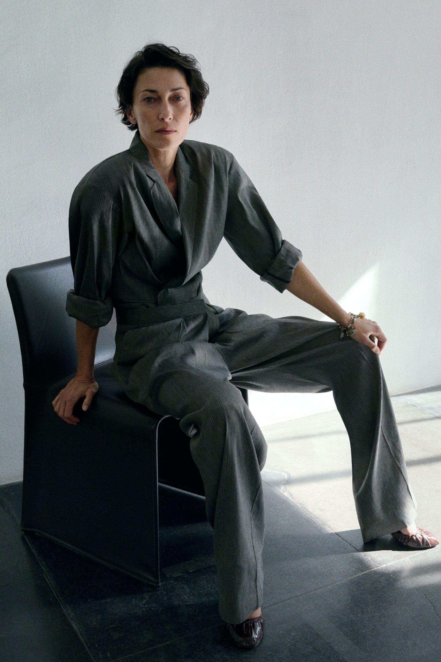 Grey pinstriped jumpsuit with a wrapped shawl lapel and self belt. Charm bracelet stacked at one wrist. Crushed metallic ballet flats, one toe catching the light. Sun on the office floor after everyone else has gone to lunch.