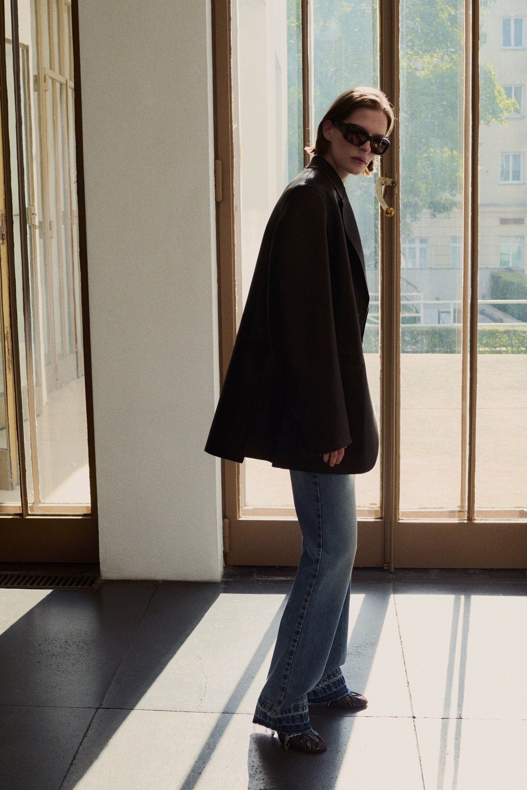Black wraparound frames with a sharp upward slant. Oversized black blazer worn cape-like, sleeves hanging past the hands. Pale blue bootcut denim with a slightly frayed hem break. Dark ruched leather flats, pinched tight across the toe. Sunlit apartment viewing, keys still warm from the broker's hand.