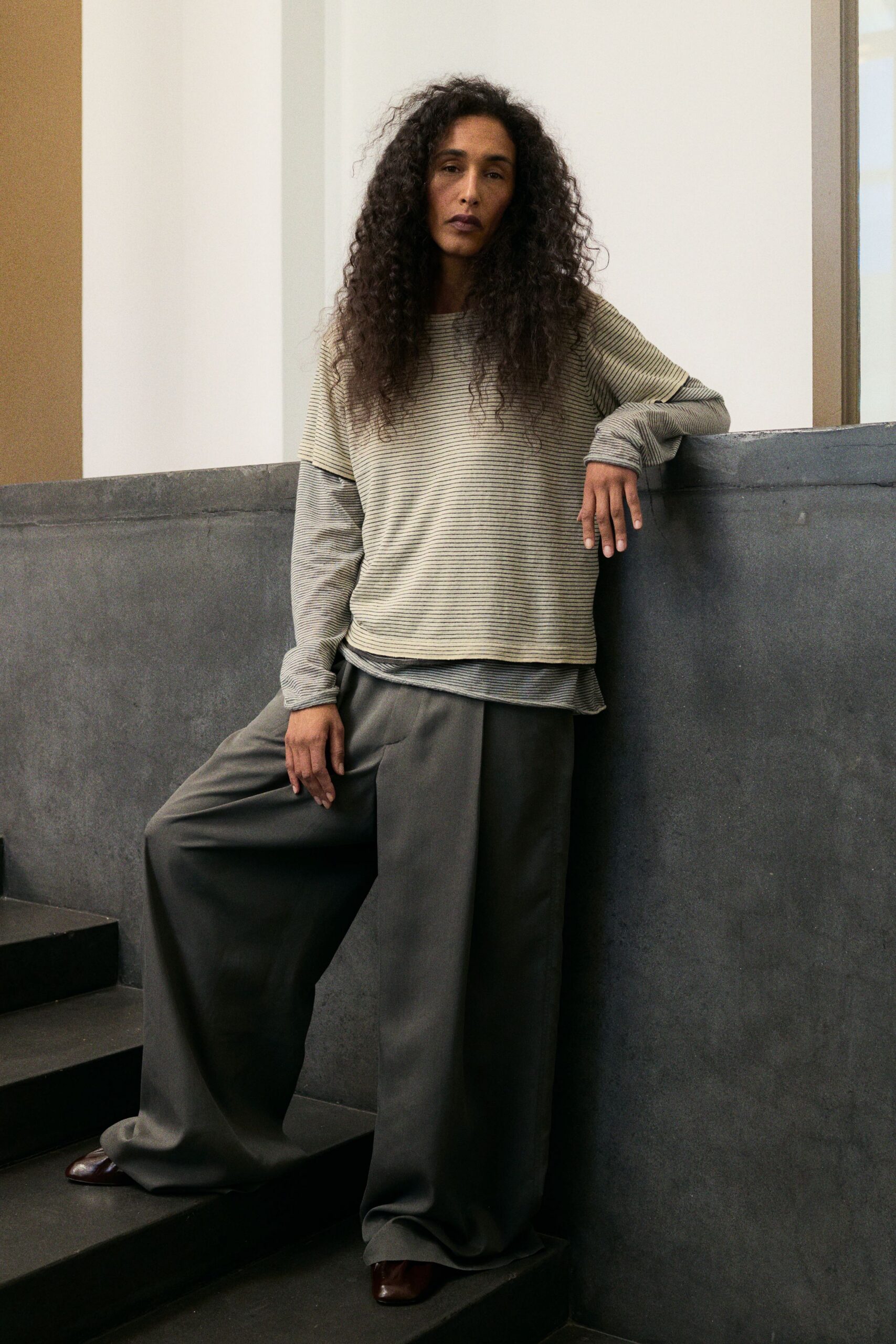 Oatmeal and charcoal striped jersey top with dropped shoulders. Finer striped underlayer, cuffs stretched long over the hands. Charcoal wide-leg trousers with a deep front pleat breaking softly. Dark oxblood square-toe shoes peeking from the hem. Climbing the museum stairs before the building opens to the public.