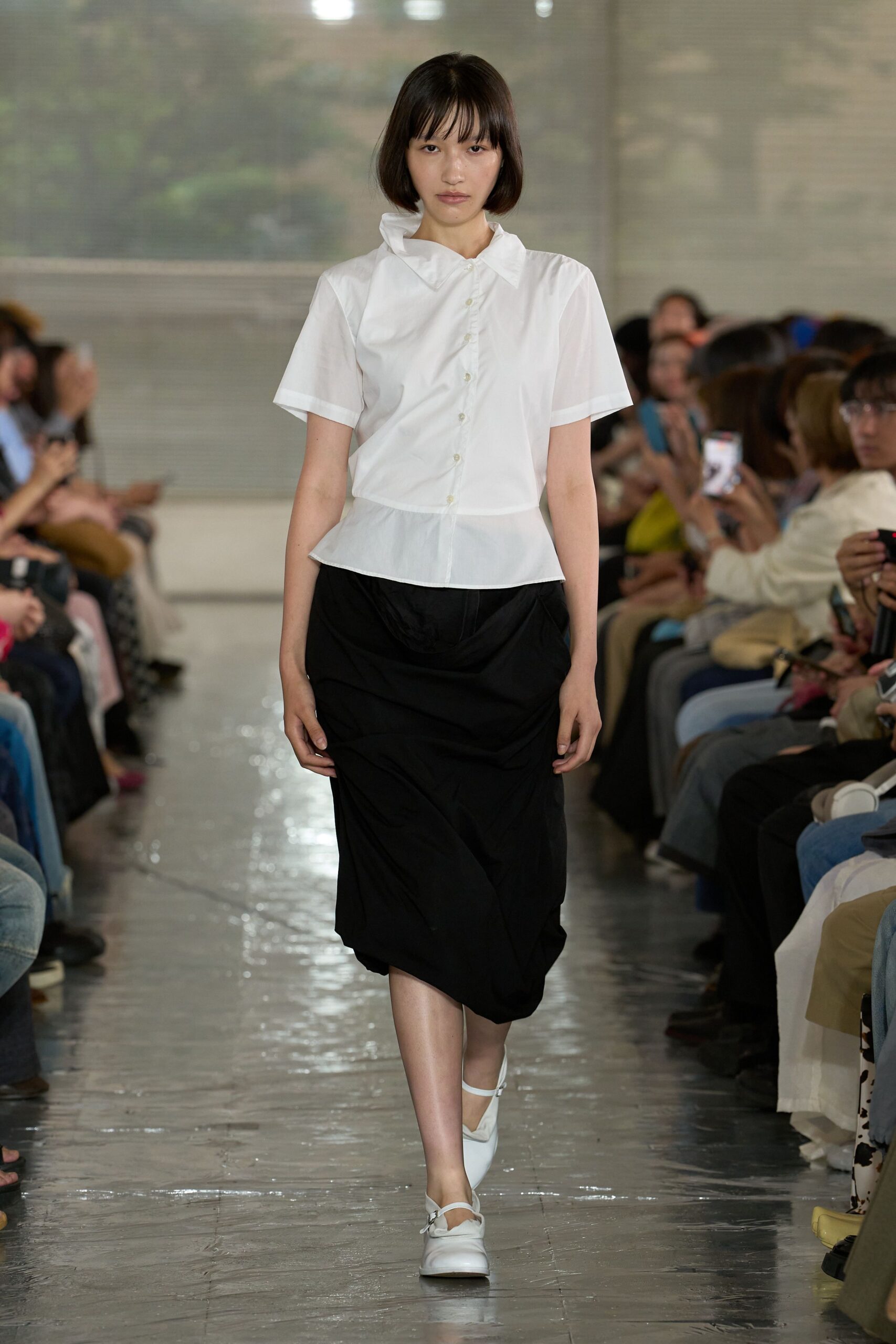 White poplin short-sleeve shirt with a skewed collar and flared peplum hem, black skirt draped low in crisp technical fabric, white strap shoes with squared toes. Like stepping through a museum lobby before the rain lets up.