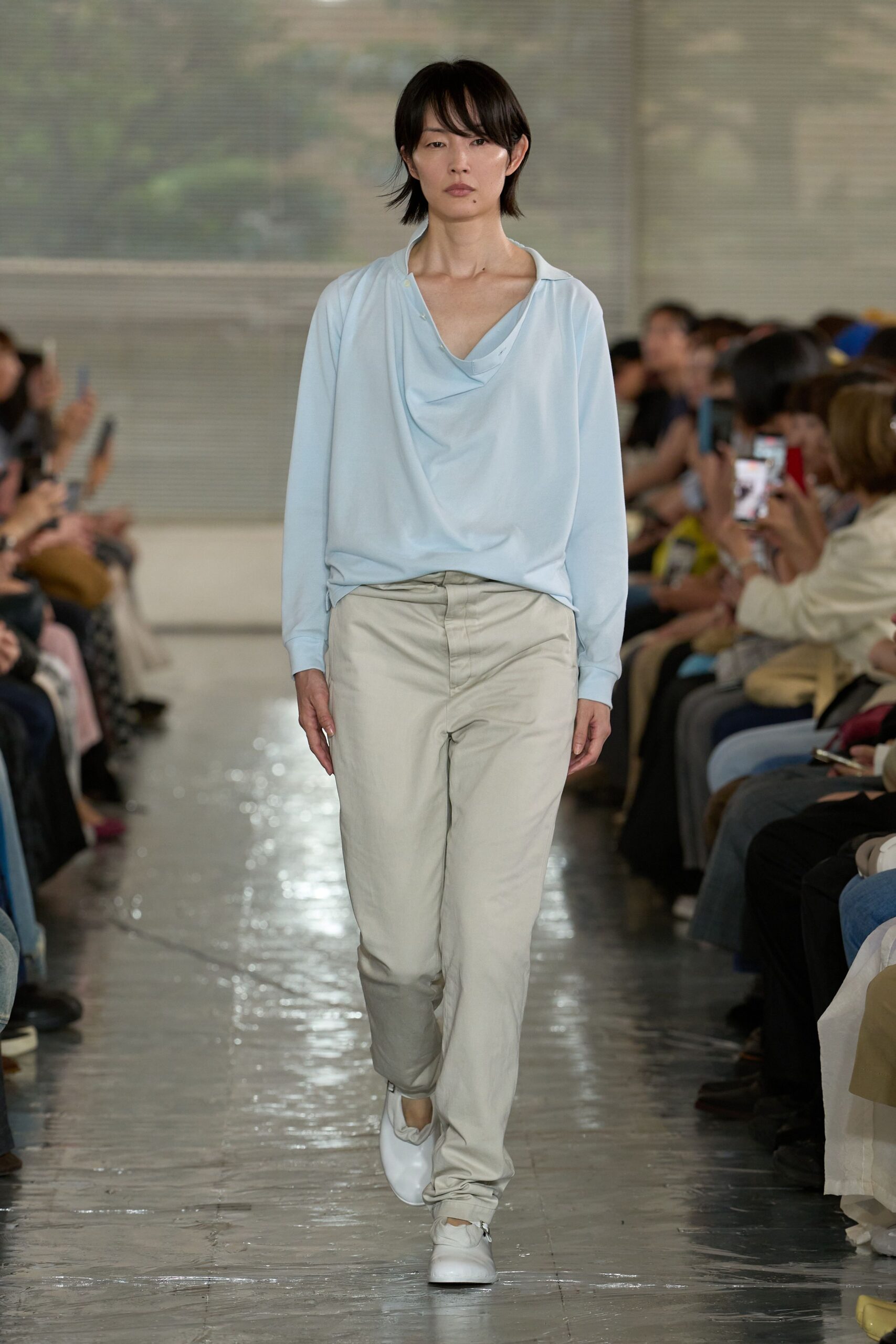 Powder-blue blouse in fluid jersey with a draped cowl and one undone collar button, chalk trousers in washed cotton with a soft break at the shoe, white leather slip-ons. Early flight, gate coffee gone cold.