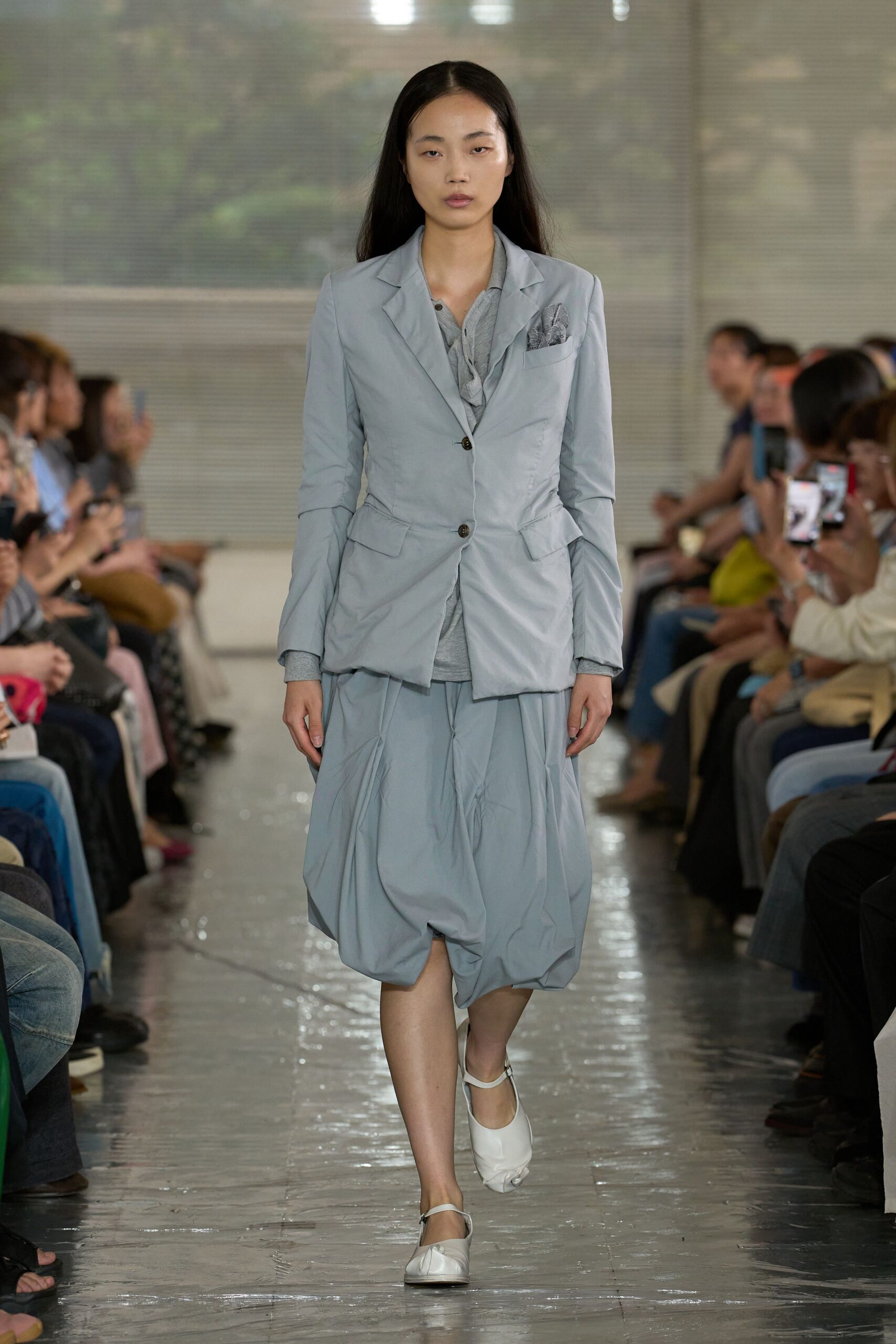 Powder-blue blazer in crinkled poplin, pinched at the waist with dark horn buttons over a heather-grey jersey polo. Matching ballooned skirt, white Mary Janes. Like arriving early to a conservatory recital in damp heat.