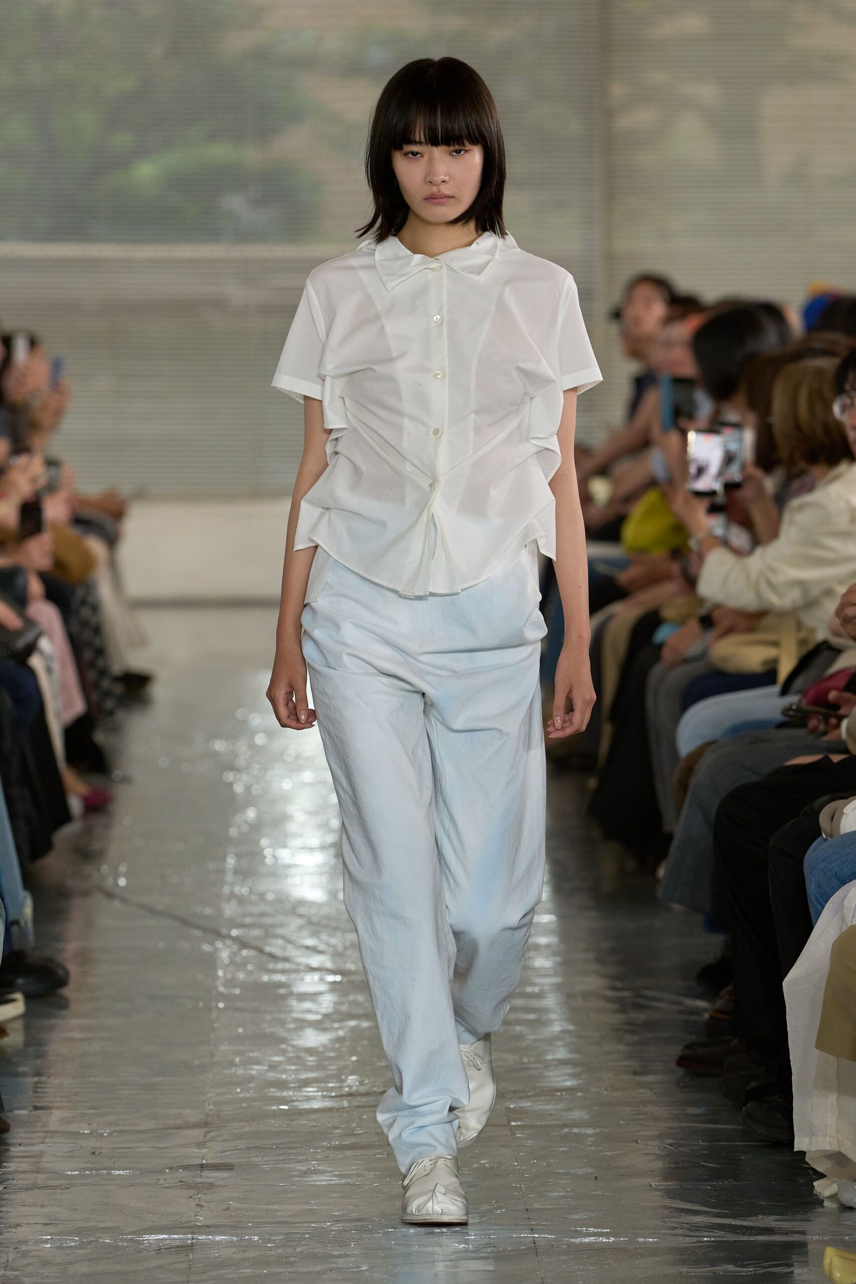 White cotton shirt with a tiny bow collar and pinched ruching that puckers down both sides. Pale wide-leg trousers in washed twill, pooling over square-toe white shoes. Fluorescent office lobby, after the cleaning crew.
