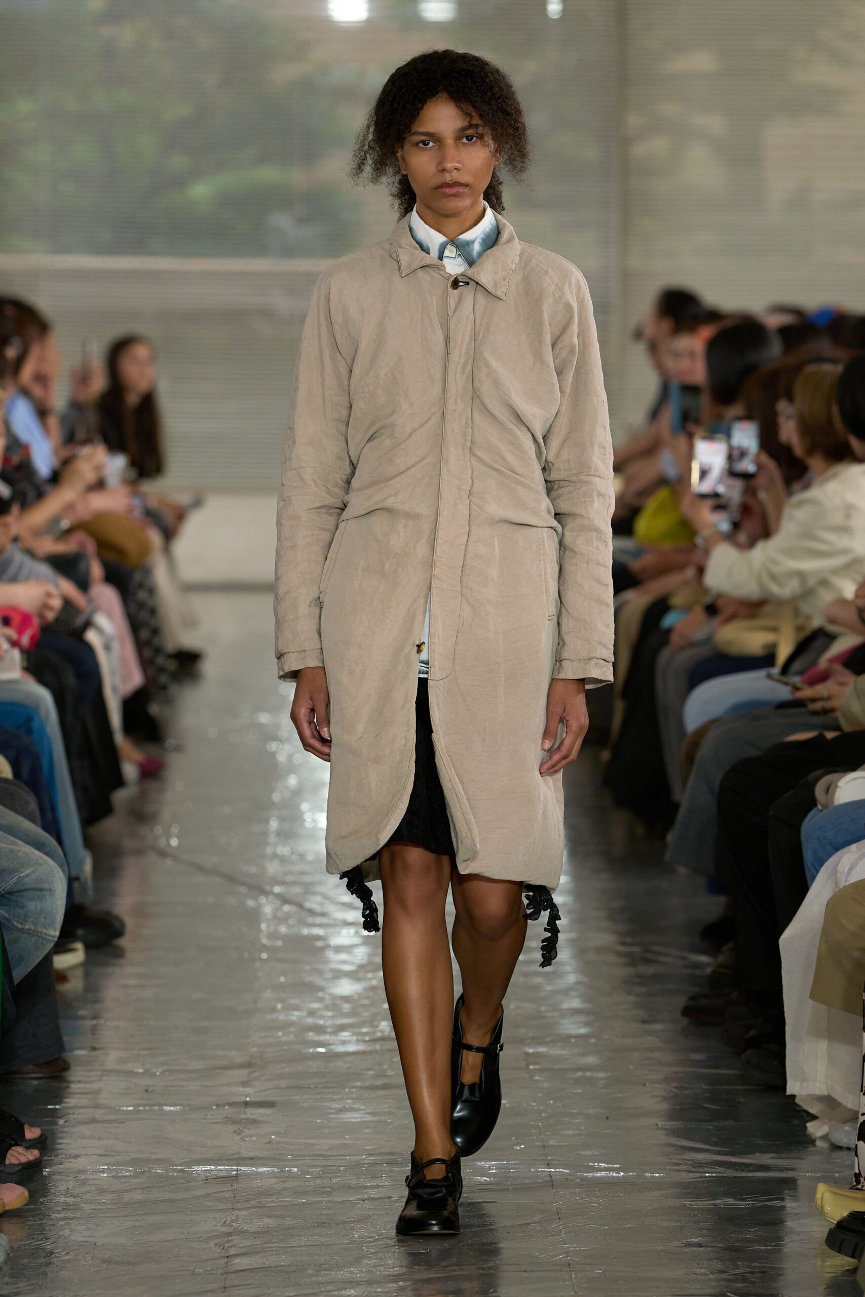 Crinkled beige coat with a curved front hem and hidden placket over a pale blue shirt washed darker at the collar. Black skirt peeking below, black Mary Jane flats. Like arriving early to a damp civic hall.