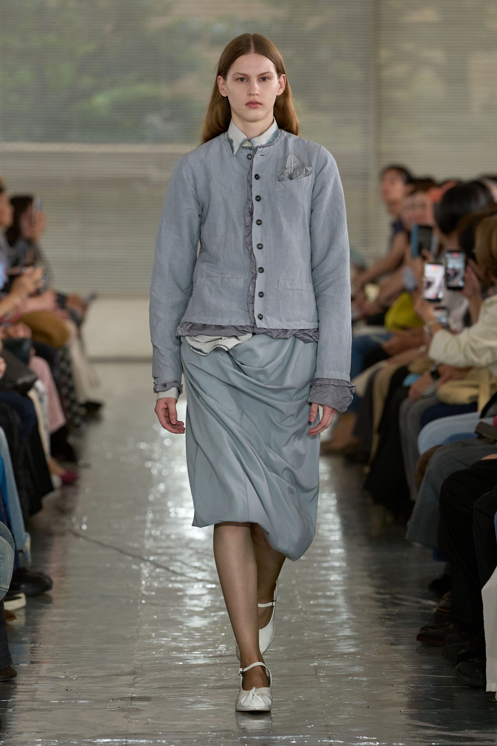 Powder-blue shirt jacket in washed cotton, black buttons marching over a crinkled placket. Pale blue draped skirt with a twisted fold at the hip, white strap flats. Like stepping out after rain with receipts in your pocket.