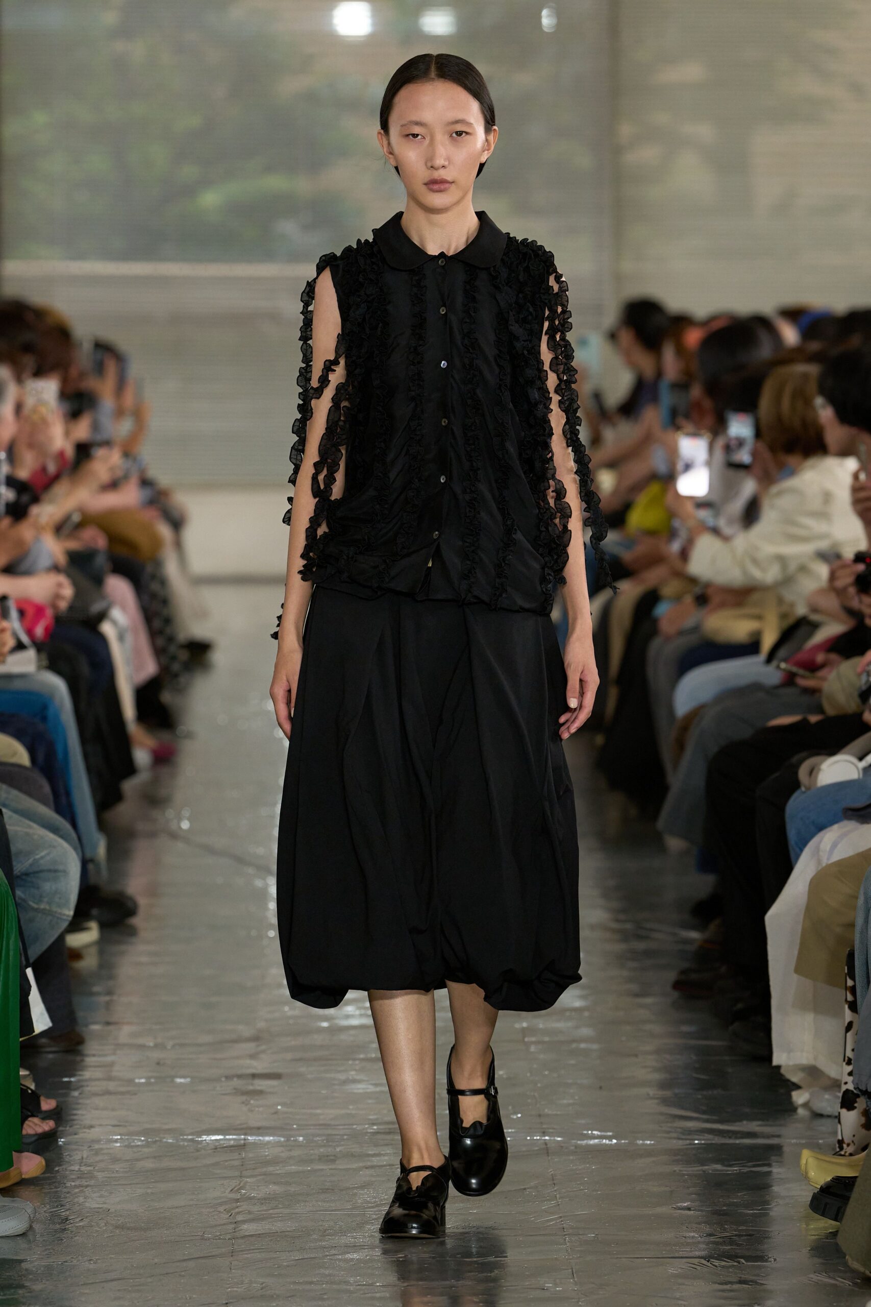 Black sleeveless blouse with a rounded collar and sheer ruffles running in vertical strips, the arm openings cut open to the shoulder. Black balloon midi skirt, glossy Mary Janes. Like waiting in a museum lobby after the rain lets up.