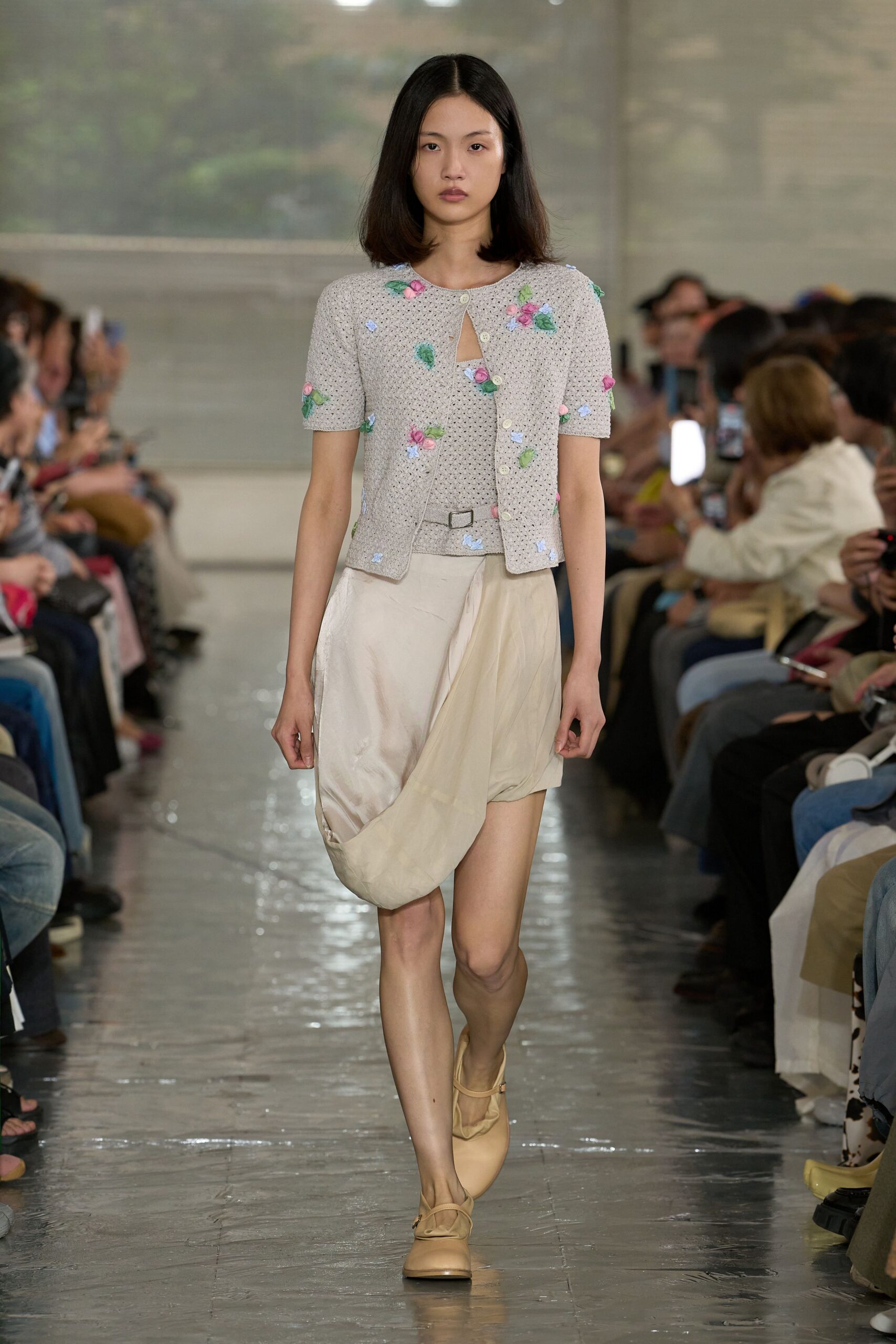 Openwork knit cardigan dotted with tiny ribbon rosettes and a low buckle tab at the waist. Ivory satin skirt draped into a pouching tulip hem. Butter leather Mary Janes. Hotel corridor at dawn, carrying yesterday’s corsage.