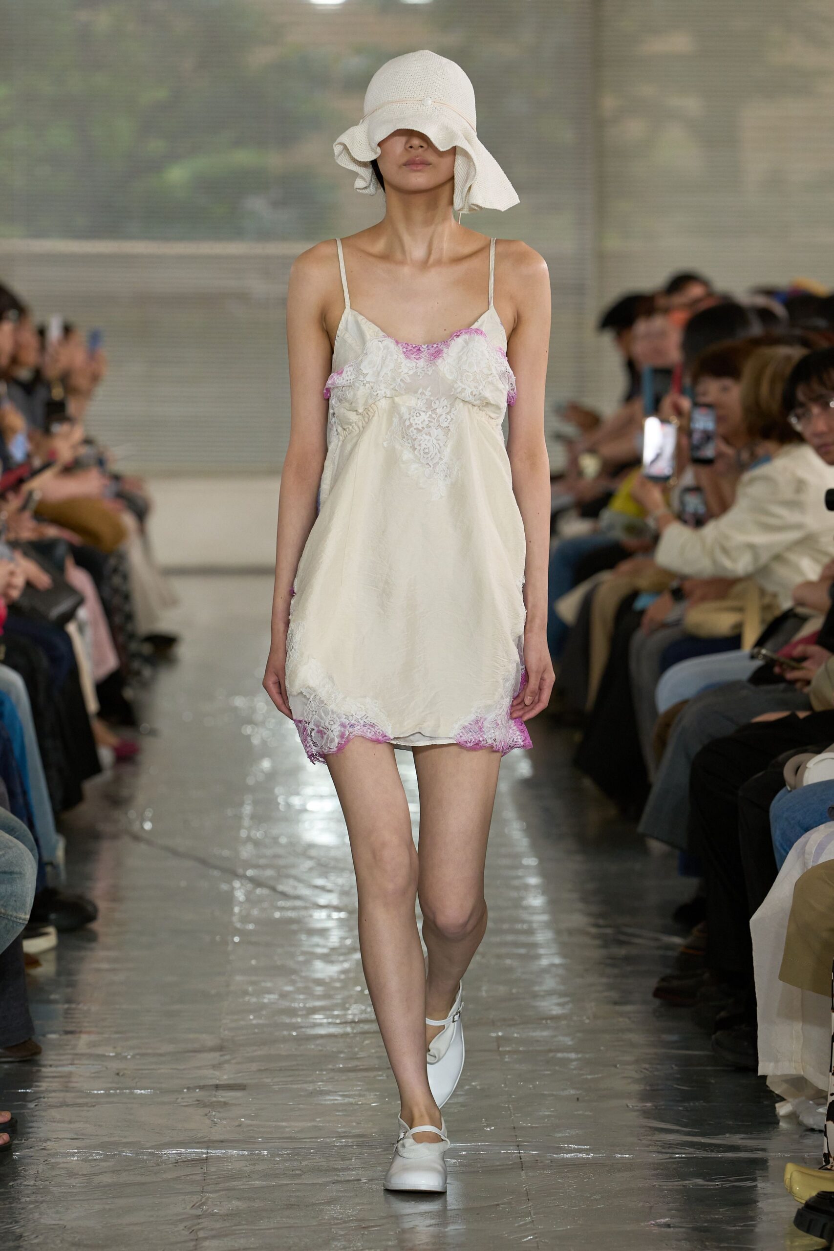 Cream slip dress in washed satin, trimmed with lilac eyelash lace that dips into a V at the bust. White cloche hat in open knit with a rippled brim, white Mary Janes. Early checkout, hotel corridor still smelling of pool chlorine.