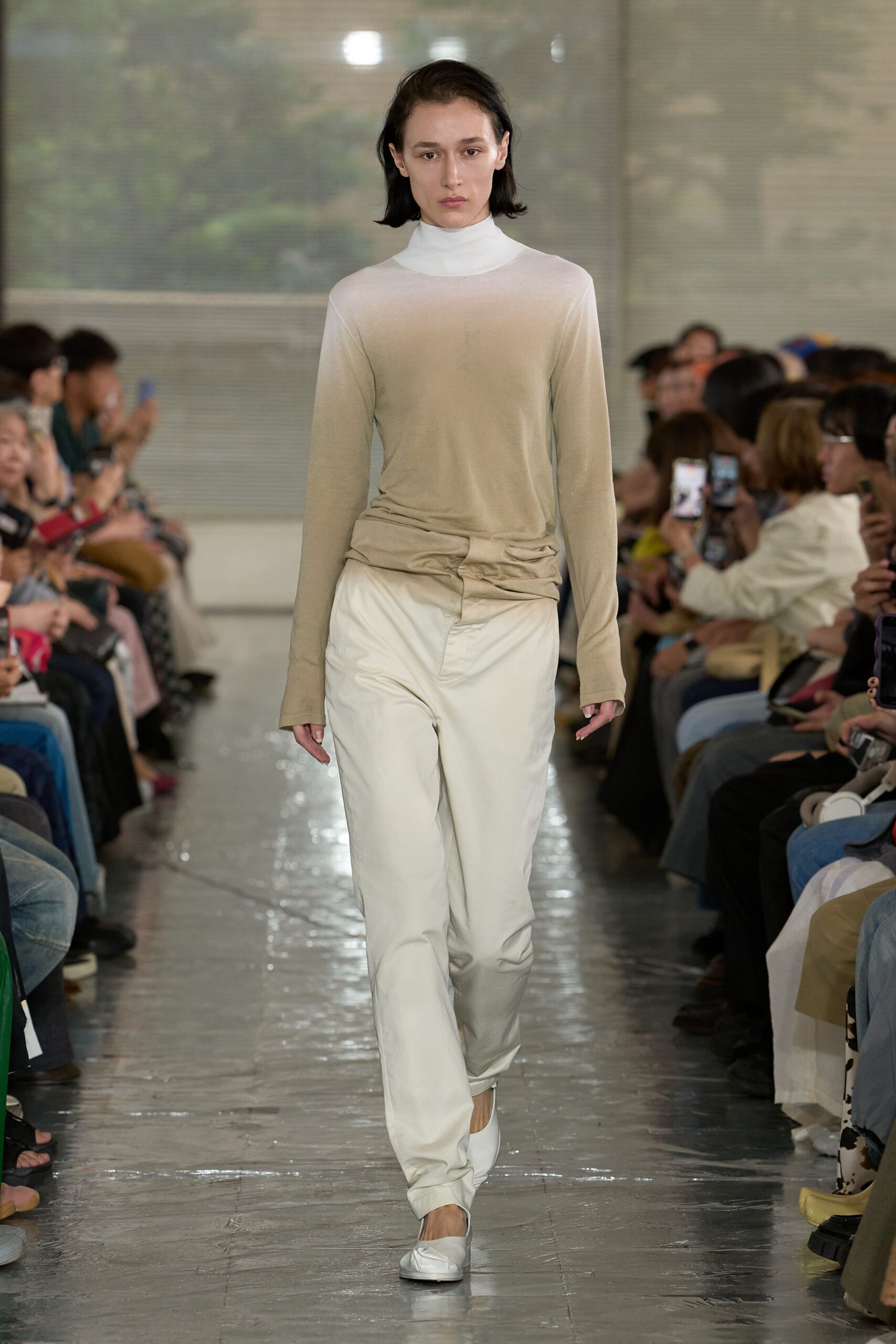 Sheer putty jersey top over a white mock-neck shell, the hem twisted into a soft knot at the hip. White trousers with a pressed front crease, white square-toe flats. Early morning in a quiet terminal café.