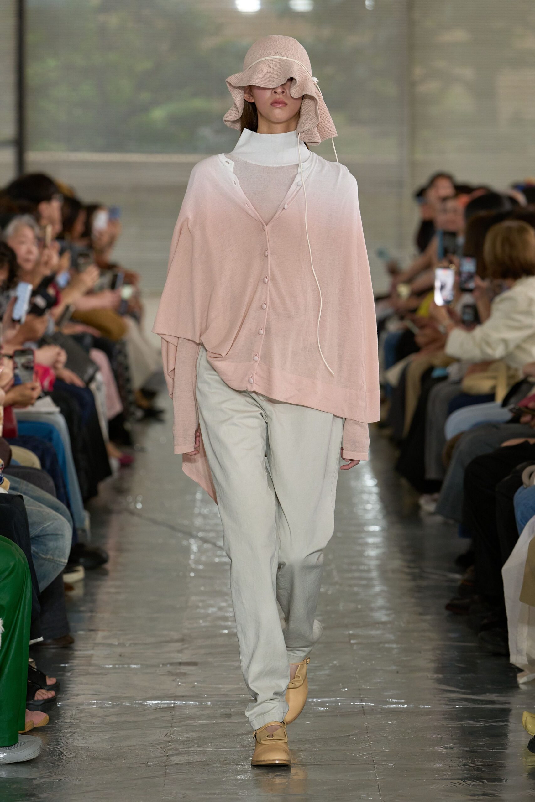 Blush waffle-knit sun hat with a drooping brim, sheer petal-pink cardigan hanging askew over a white funnel-neck layer, chalk trousers with a loose break, beige leather shoes. Crossing a museum atrium before it opens.