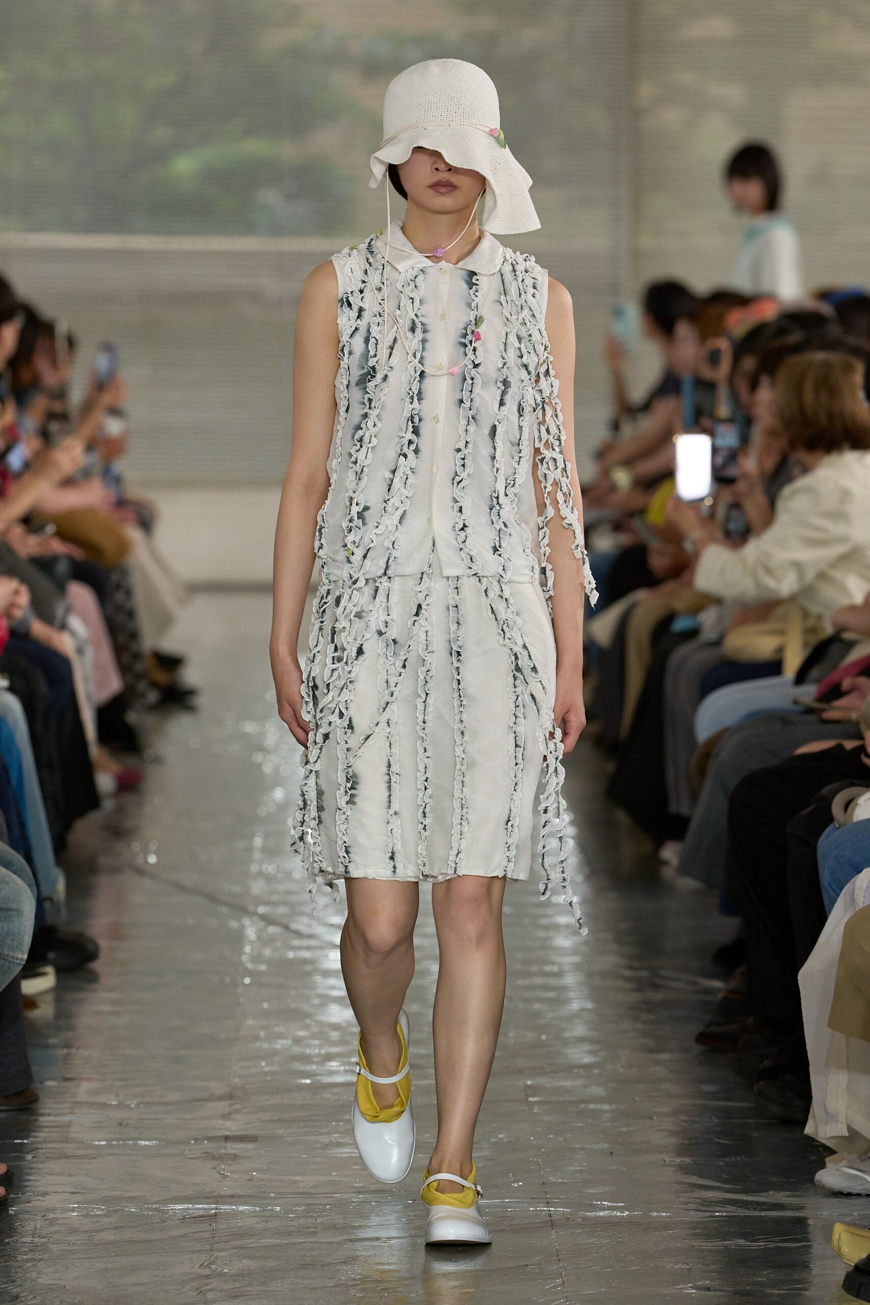 White open-weave sun hat with a drooping brim, sheer sleeveless blouse traced in narrow ruffled strips, matching knee-length skirt with the same fluttering seams, white Mary Janes lined in citron. A conservatory walkway just after watering.