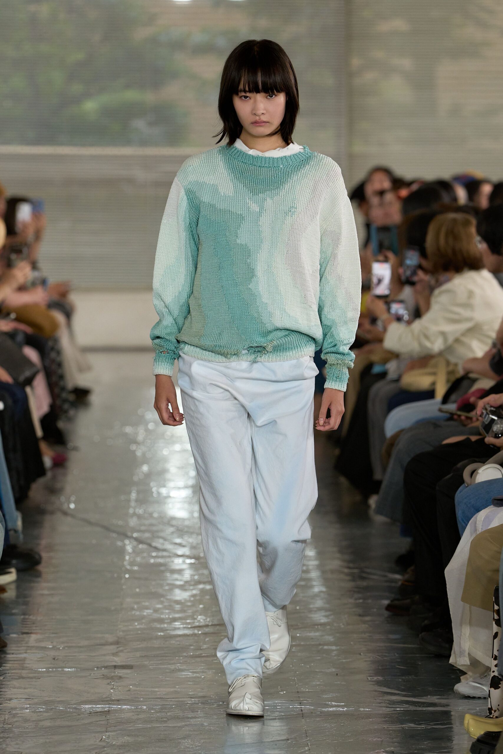 Mint-and-chalk sweater in a loose open knit, the hem visibly nibbled away at center front. White crewneck peeking at the collar, white trousers with a soft crushed finish, white square-toe shoes. Like stepping out after rain at a seaside motel.