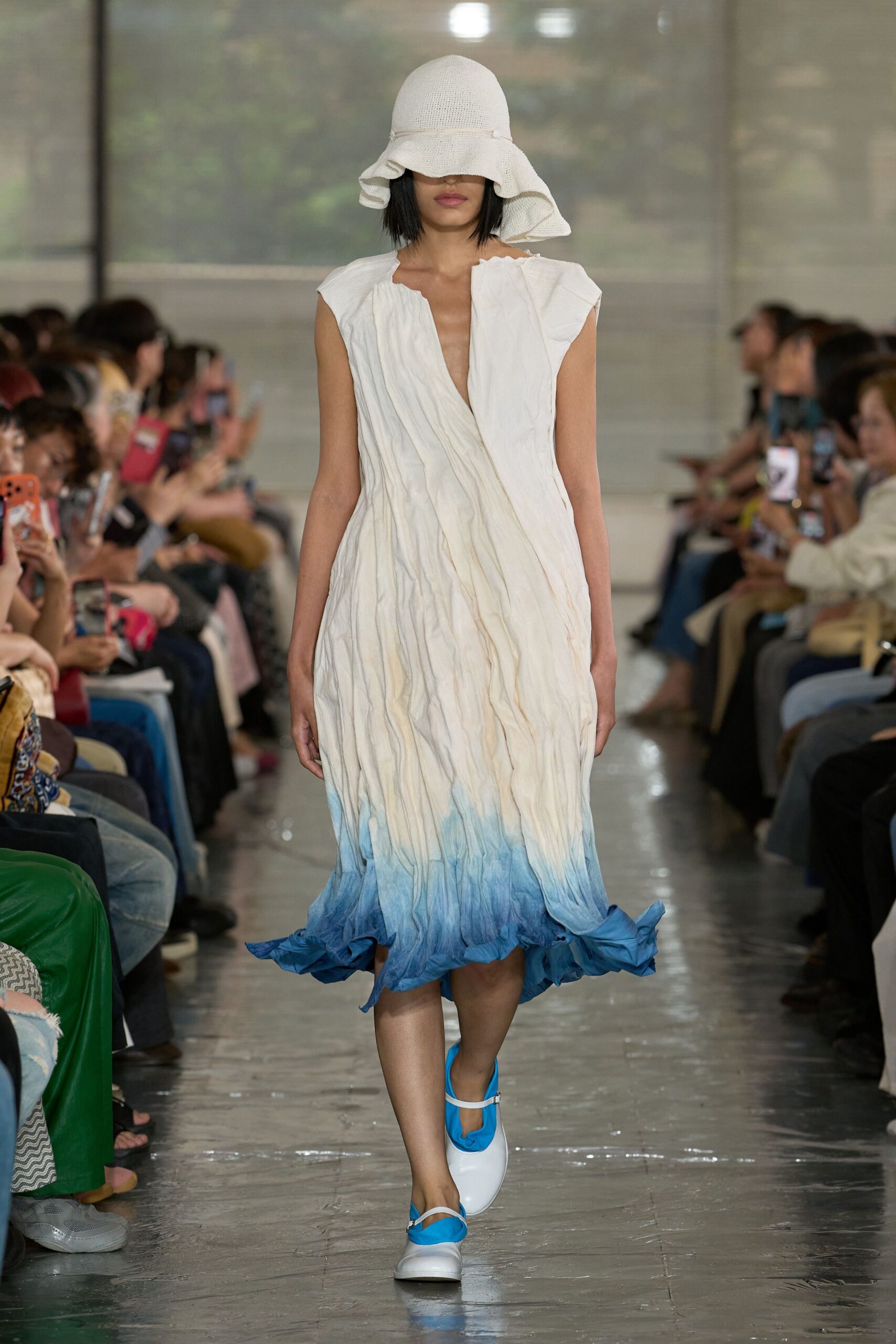 Floppy white open-weave sunhat with a wavering brim. Sleeveless white dress in crinkled cloth, wrapped at the bodice and washed to cobalt at the ruffled hem. White Mary Janes with bright blue toes. Seaside greenhouse, just after rain.