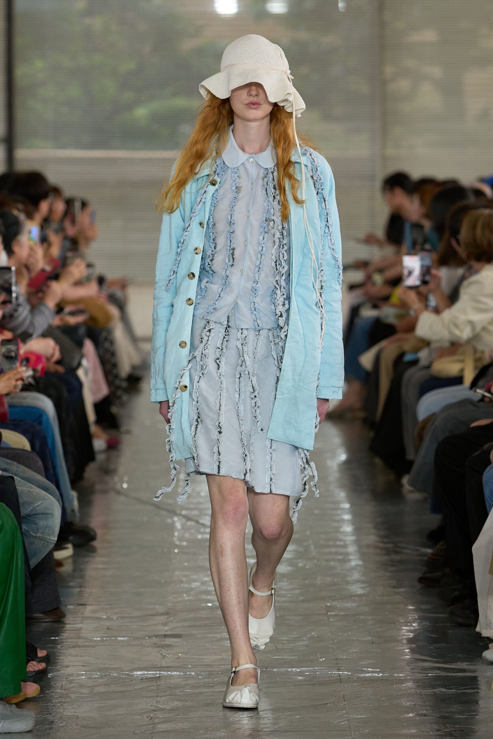 Cream open-knit sun hat with a drooping brim and long tie cords. Pale blue coat with horn buttons over a sheer blouse and matching skirt, both traced in fluttering ruffle strips. White Mary Janes. Like stepping out after rain near the conservatory.