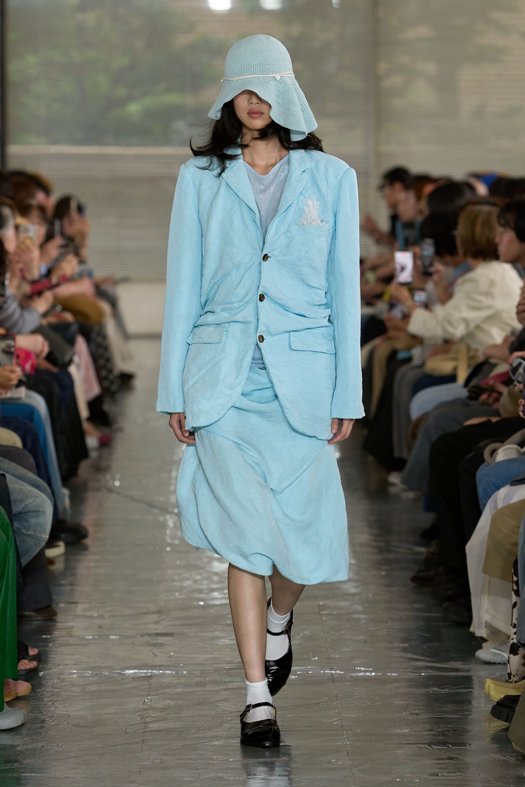 Powder-blue open-weave cloche with a cord circling the brim, crinkled sky-blue blazer with dark horn buttons, sheer ribbed knit top, softly ruched matching skirt, white socks and black patent Mary Janes. Like leaving a seaside matinee into wind.